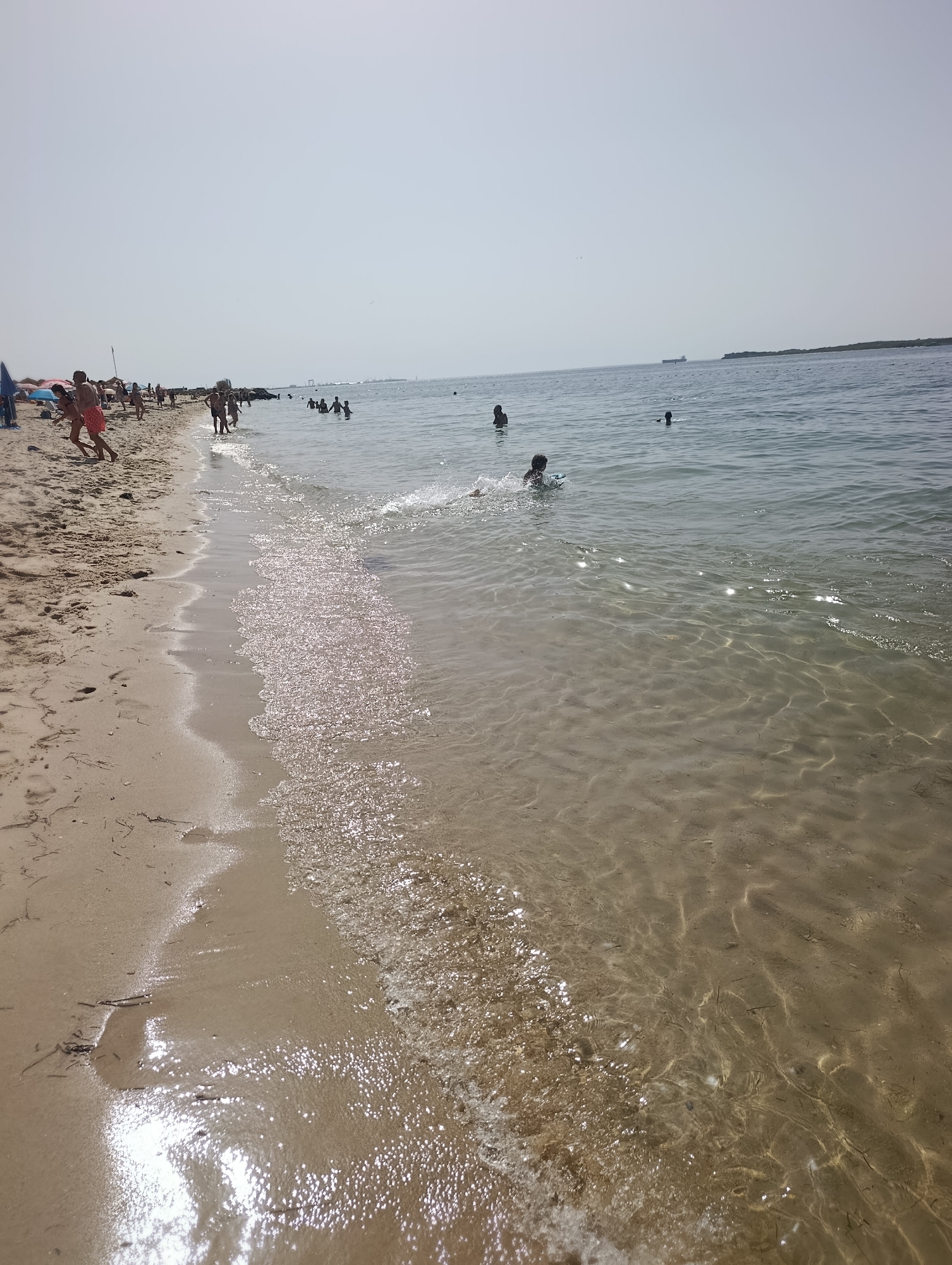A wide-angle, eye-level view of a sandy beach on a clear, sunny day. The foreground shows the wet sand where waves are gently lapping. Several people are visible on the beach and in the shallow water. On the left, a group of people are gathered, some wading, others standing. Further out in the water, several individuals are swimming or playing. In the distance, the sea stretches to the horizon under a pale sky, with a large ship visible far off. The overall atmosphere is one of a relaxed, casual day at the beach. The location is Praia da Figueirinha in Setúbal, Portugal.