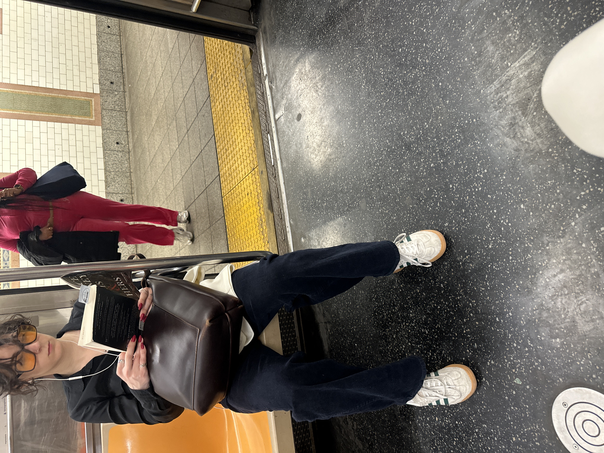 The image captures a scene inside a subway train, looking out onto a station platform. The train door is open, revealing the platform beyond.
In the foreground, a person is seated on an orange subway bench. They have short, curly dark hair, are wearing a black long-sleeved shirt, dark blue wide-leg trousers, and white sneakers with green stripes and tan gum soles. They are wearing orange-tinted rectangular sunglasses and wired white earphones. Their fingernails are painted red, and they wear rings on several fingers. This individual is engrossed in reading a black-covered paperback book, which they hold in both hands. A large, dark brown leather bag rests beside them on the seat.
Outside the train, on the platform, another person is partially visible. They are wearing a bright pink tracksuit with white side stripes and white sneakers. They are holding a dark bag and appear to be standing or walking on the platform.
The subway car features a speckled dark gray floor and orange seating. The platform outside has white tiled walls with a decorative horizontal band of green and brown tiles. The platform floor is a mix of gray square tiles and prominent yellow tactile paving along the edge.
The activity depicted is commuting, with one person reading on the train and another on the platform. The style of the main subject, with vintage-inspired sunglasses and wired earphones, suggests a particular aesthetic. The only clearly visible text is "HERS" on the spine or cover of the book being read. Given the context, this scene is set in the New York City subway system.