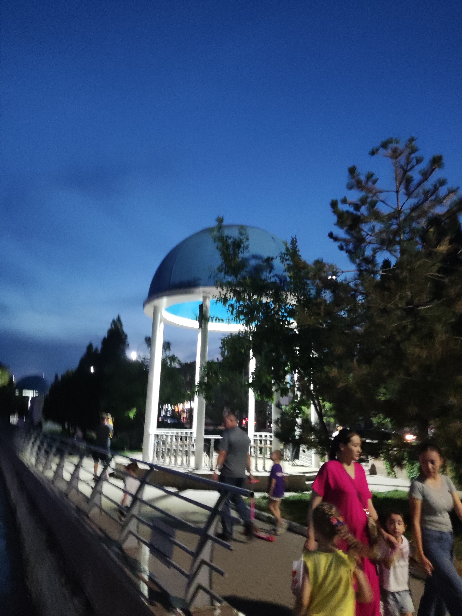 This is a medium shot photograph taken at dusk in a park in Shymkent, Kazakhstan. The sky is a deep blue, indicating the transition from day to night. In the center of the image, a white gazebo with a blue dome stands prominently. It is illuminated from within, casting a soft glow.
Several people are visible walking along a paved pathway that borders a body of water on the left side of the frame, suggested by a dark, reflective surface. A metal railing runs along the pathway.
In the foreground, a woman in a bright pink dress is walking with two children. One child, in a yellow shirt, is in front of her, and another child, wearing a white shirt and shorts, is beside her. To the right, another woman in a grey t-shirt and jeans is walking. Further in the background, a man in a grey shirt and jeans is walking. A young child in a purple shirt and white shorts is also visible.
The overall atmosphere appears to be peaceful, with people enjoying an evening stroll in the park. The presence of the gazebo and the well-maintained pathway suggests a public recreational area. The lighting suggests it is after sunset, but before complete darkness. No visible text is present in the image.