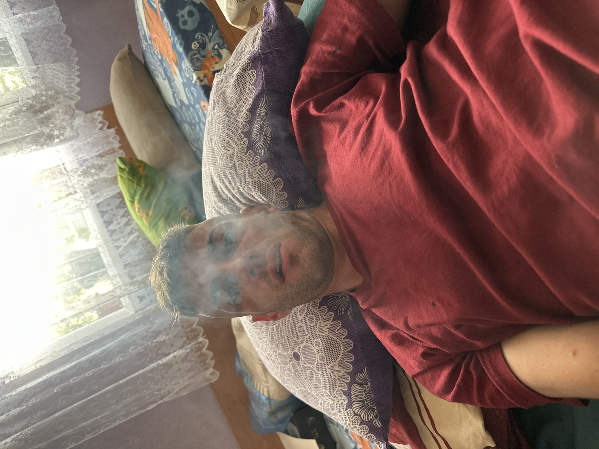 A man is lying down on a bed, with smoke visible around his face. He is wearing a red t-shirt. The bed is covered with several pillows, some of which have intricate patterns. Behind the bed, there is a window with sheer white curtains, through which natural light is filtering in. The overall setting appears to be indoors, possibly in a bedroom. The image does not provide specific details about the location in Cherven Breg, Bulgaria.