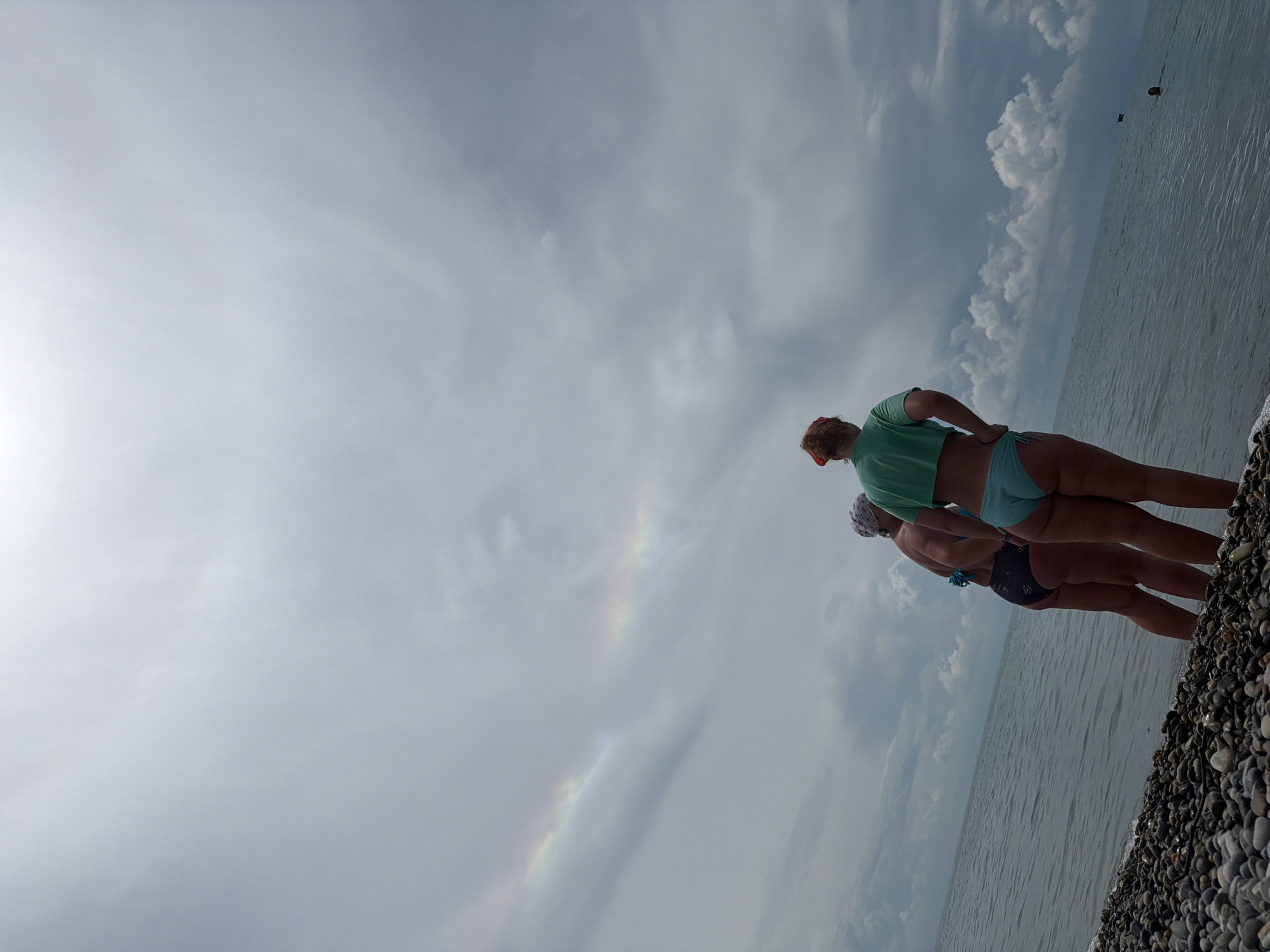 Two women stand on a pebble beach in Sochi, Russia, facing the sea under a cloudy sky. A faint rainbow is visible in the sky above them. The woman on the left wears a dark bikini, and the woman on the right wears a light blue bikini and a green crop top, with a red bandana on her head. The sea is calm, and the sky is filled with dramatic clouds.