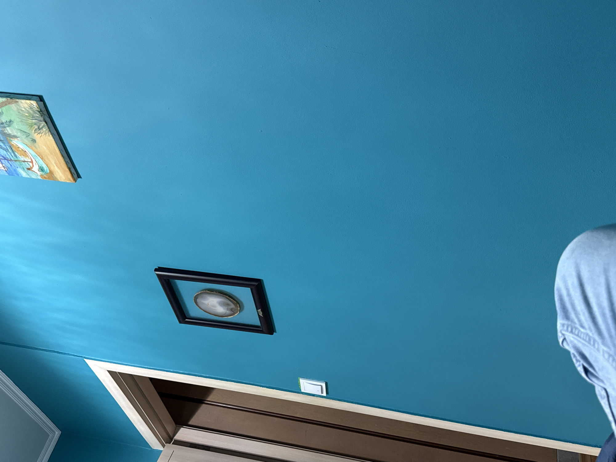 The image shows an indoor scene with a predominantly blue ceiling and walls. In the upper left corner, a framed painting or artwork is visible. A square frame containing a circular object, possibly a decorative plate or stone, hangs on the ceiling. Below the frame is a doorway with a wooden door and white trim. A light switch is located above the door frame. Part of a person's blue clothing appears on the right side of the image. It is not possible to determine the location in Varna, Bulgaria from the image.