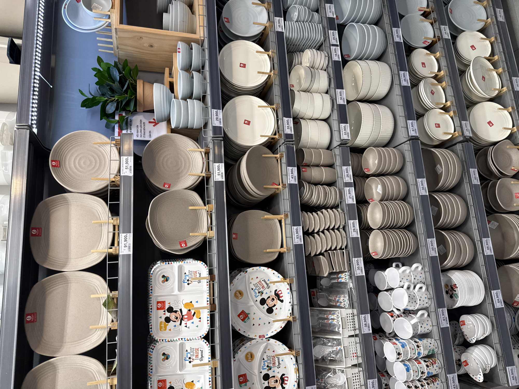 The image shows a retail store display of tableware in Nha Trang, Vietnam. The shelves are filled with various plates, bowls, and cups, mostly in neutral and pastel colors, with some featuring Disney characters like Mickey Mouse. Prices are displayed on tags in Vietnamese Dong. The store appears to be well-organized, with items neatly stacked and arranged on metal shelving units. The overall lighting suggests an indoor environment, likely a department store or a specialized kitchenware shop. No people are visible in the image.