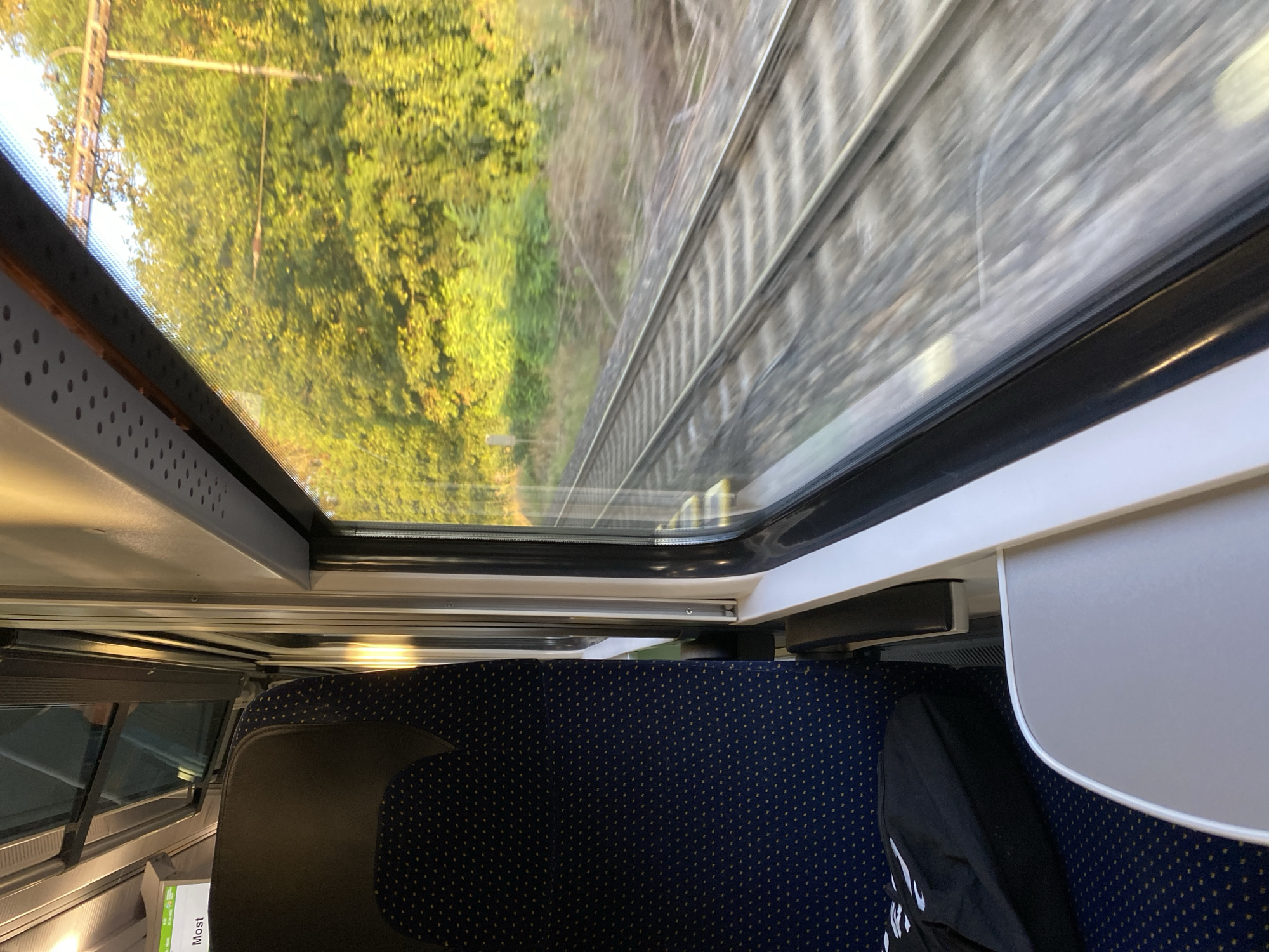 The image shows the interior of a train carriage, looking out the window. A dark blue seat with small yellow dots is visible on the left, along with a black bag on the floor. A fold-down table is in the foreground. Outside the window, train tracks are visible, with trees and greenery in the background. The motion blur indicates the train is in transit. The scene is captured during daylight, with sunlight filtering through the trees, creating a warm, golden hue. The train is likely traveling through a rural or suburban area, given the dense foliage. While the image does not explicitly state the location, the presence of a train and the surrounding landscape are consistent with travel within the Czech Republic. Bílina, Czechia, is known for its railway connections and surrounding natural beauty, making it a plausible location for such a scene.