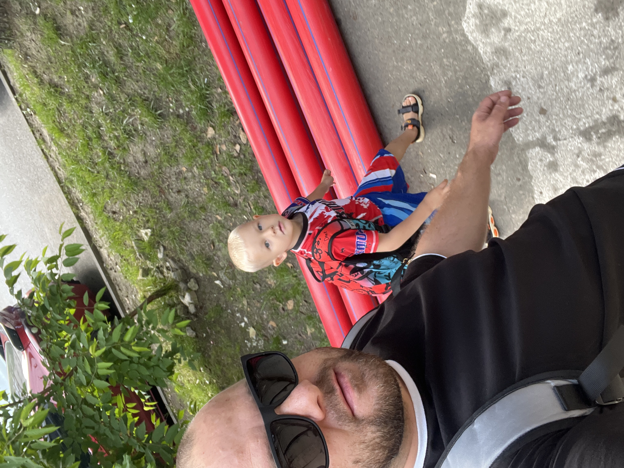 This outdoor image, captured in Sochi, Russia, features an adult male taking a selfie with a young boy on a bright, sunny day. The adult, seen from the chest up in the foreground, wears sunglasses, a black short-sleeved shirt with white trim, and a black and grey backpack strap. He has a beard or stubble.
In the middle ground, a young boy with blond hair looks directly at the camera. He is wearing a colorful t-shirt with red, blue, and black patterns, where partial text like "Don't" and "Amuse" is visible. He also has on red, white, and blue striped shorts and sandals. The boy is seated on a stack of several long, bright red pipes, which have thin blue lines running along their length. These pipes rest on a paved surface next to a strip of patchy grass and dirt.
The background reveals green foliage from bushes or trees, and a portion of a red car is visible behind the greenery on the left. The ground consists of both asphalt or pavement and areas of sparse grass and dirt. The overall scene suggests a relaxed, casual outing in an urban or semi-urban environment.