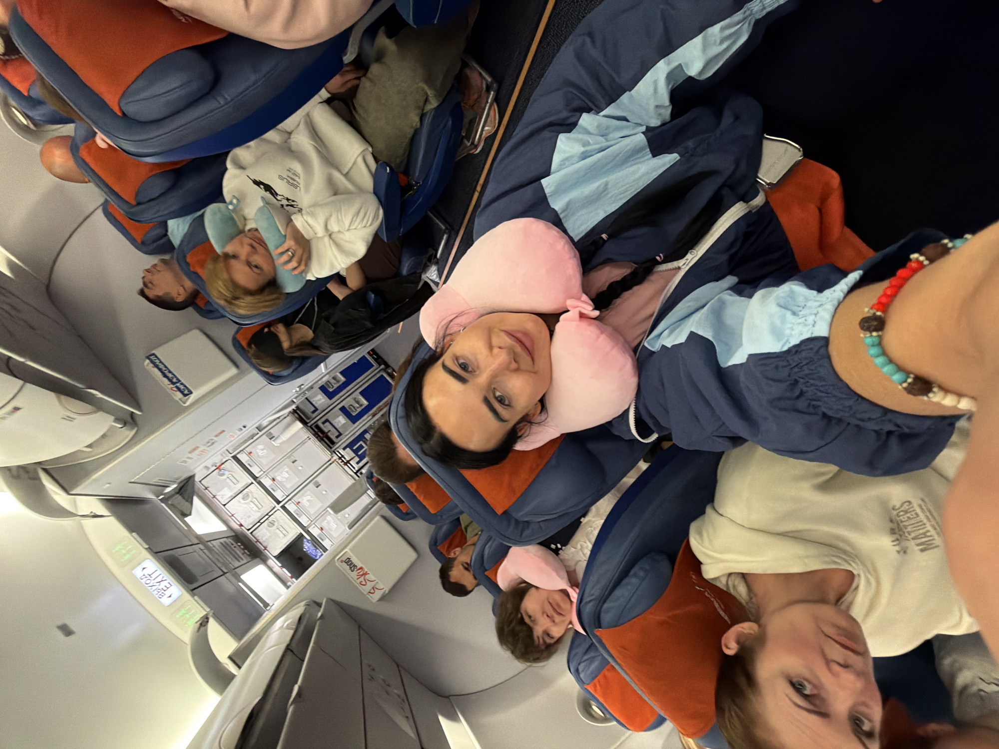 The image shows the interior of an airplane cabin, likely a commercial flight. Multiple passengers are seated in blue and orange airplane seats, some of whom appear to be sleeping or resting with neck pillows. The overhead compartments and lighting fixtures are visible, along with an "EXIT" sign in English and Russian. One passenger in the foreground is taking a selfie, smiling at the camera and wearing a neck pillow. Another passenger to her left is wearing a sleep mask. The overall scene depicts the typical environment of an airplane during a flight. The location is not in the city of Saint Petersburg, Russia.