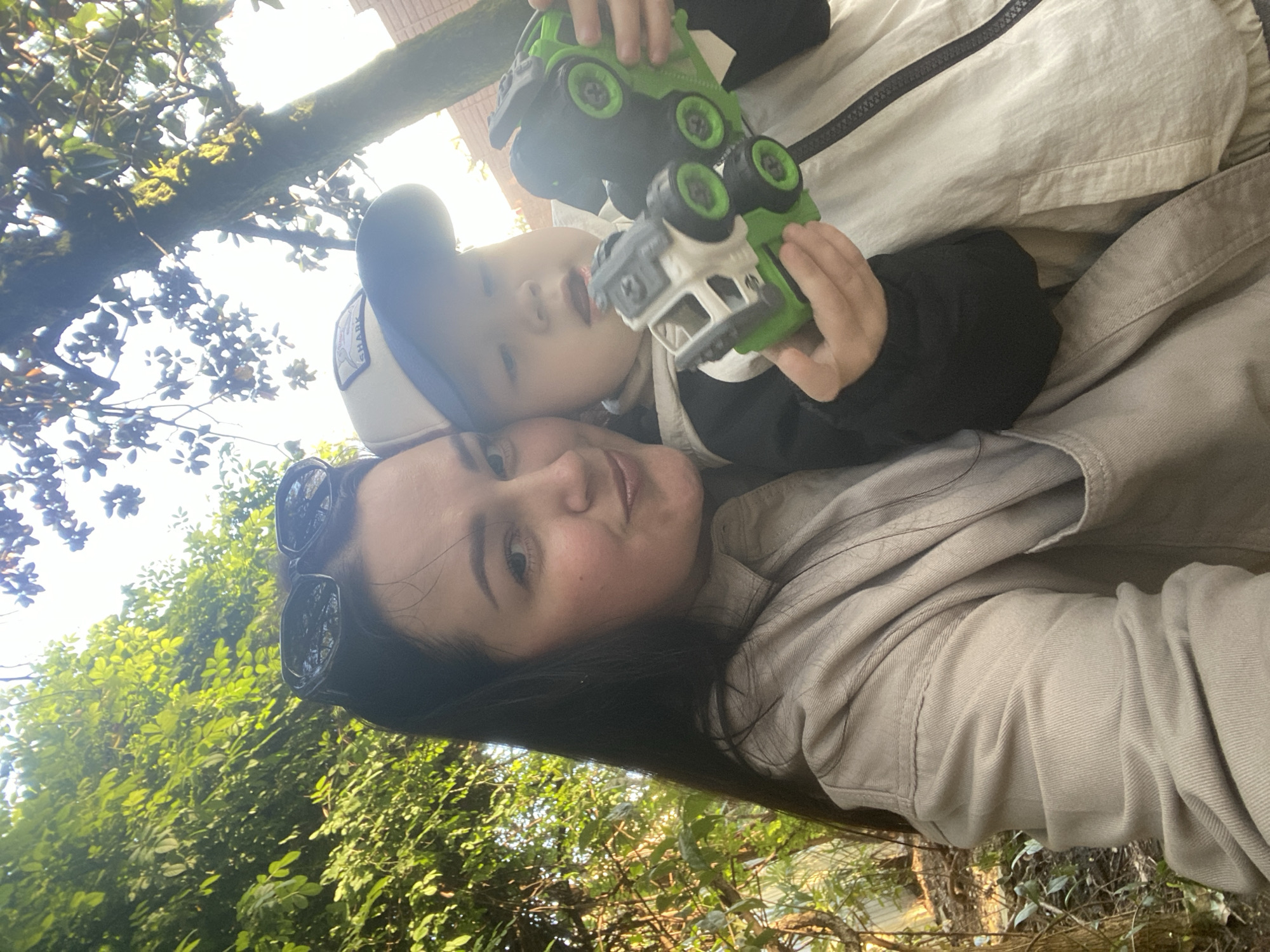 The image captures a selfie featuring a woman and a young child, taken outdoors in a lush, green environment. The photo is rotated 90 degrees clockwise, giving it a tilted perspective.
**Subjects:** The primary subjects are a woman with dark hair and a young child. The woman wears sunglasses perched on her head and a light-colored jacket. The child wears a baseball cap with a patch, a dark long-sleeved shirt, and a lighter jacket.
**Objects:** The child is holding two green and white toy trucks, one in each hand. The cap worn by the child has a visible patch.
**Scene/Setting:** The setting is outdoors, characterized by abundant, vibrant green foliage. Large tree branches, some covered in moss, are visible in the background, along with dense bushes and leaves, suggesting a park, garden, or natural area. The bright sky is visible through the canopy, indicating an open-air location.
**Activity or event taking place:** The woman appears to be taking a selfie with the child, as both are looking towards the camera. The child is holding and seemingly playing with the toy trucks.
**Notable details:**
* **Time of day:** It is daytime, evident from the bright sunlight filtering through the leaves and illuminating the scene.
* **Weather:** The weather appears sunny and clear.
* **Location cues:** The lush, green vegetation and large trees are consistent with the subtropical climate found in Sochi, Russia.
* **Emotions:** The woman has a neutral to softly smiling expression. The child's expression is calm and focused on the camera.
* **Visible text:** The word "SHARK" is visible on a patch on the child's baseball cap.