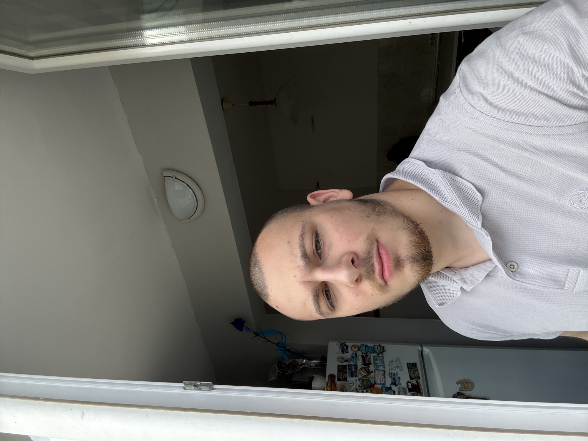 A man with short hair and a beard is looking out of an open window. He is wearing a light grey polo shirt. Behind him is a kitchen with a white refrigerator covered in magnets and stickers. In the window are a blue rose and a blue ribbon. A ceiling lamp is visible in the background. The location is Stara Zagora, Bulgaria.
