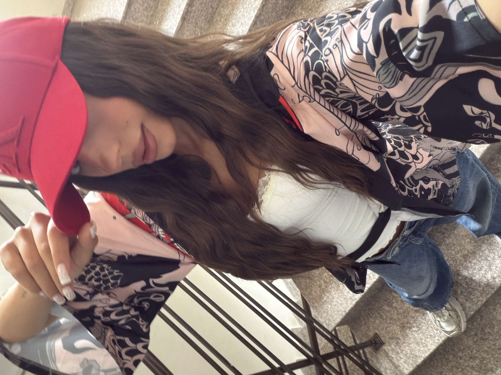 A woman is standing on a concrete staircase in Varna, Bulgaria. She is wearing a red baseball cap, a white tank top, a black and pink patterned jacket, and blue jeans. Her hair is long and brown, and she is holding her hand up to her cap. The staircase has metal railings on the left side. The image appears to be a selfie taken by the woman.