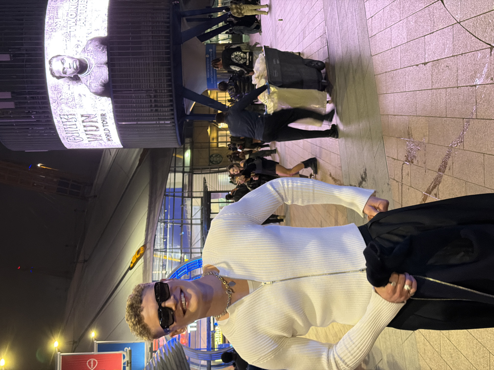 The image shows a man standing outdoors in what appears to be a public space. He is fair-skinned with short, curly blonde hair and is wearing sunglasses, a white, zipped-up sweater, and a gold chain necklace. He has a ring on one of his fingers and is holding a black jacket in his hand.
In the background, there's a crowd of people and what seems to be an entertainment venue or large building. There is a large screen displaying an advertisement or promotion featuring an image of a black male. The image also states "GIVĒON World Tour" suggesting this is near a concert venue. Given the distinctive design of the building and architectural elements, the location is very likely the O2 Arena in Greenwich, London, United Kingdom. The ground is paved with light-colored stone. The scene is lit artificially, likely at night.