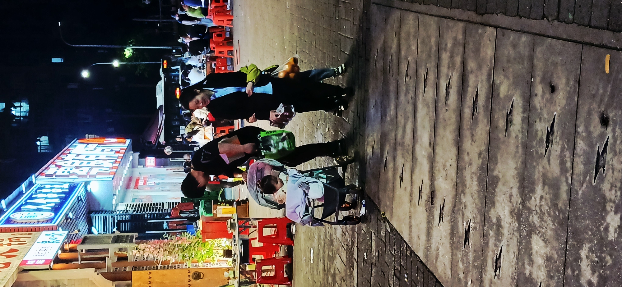 Jiulongpo District, China. An outdoor night scene shows a street-side eatery with tables and chairs. Two adults stand near the edge of the sidewalk. One pushes a stroller, while the other is partially obscured. Light poles illuminate the street. The foreground features a boardwalk with artificial damage.