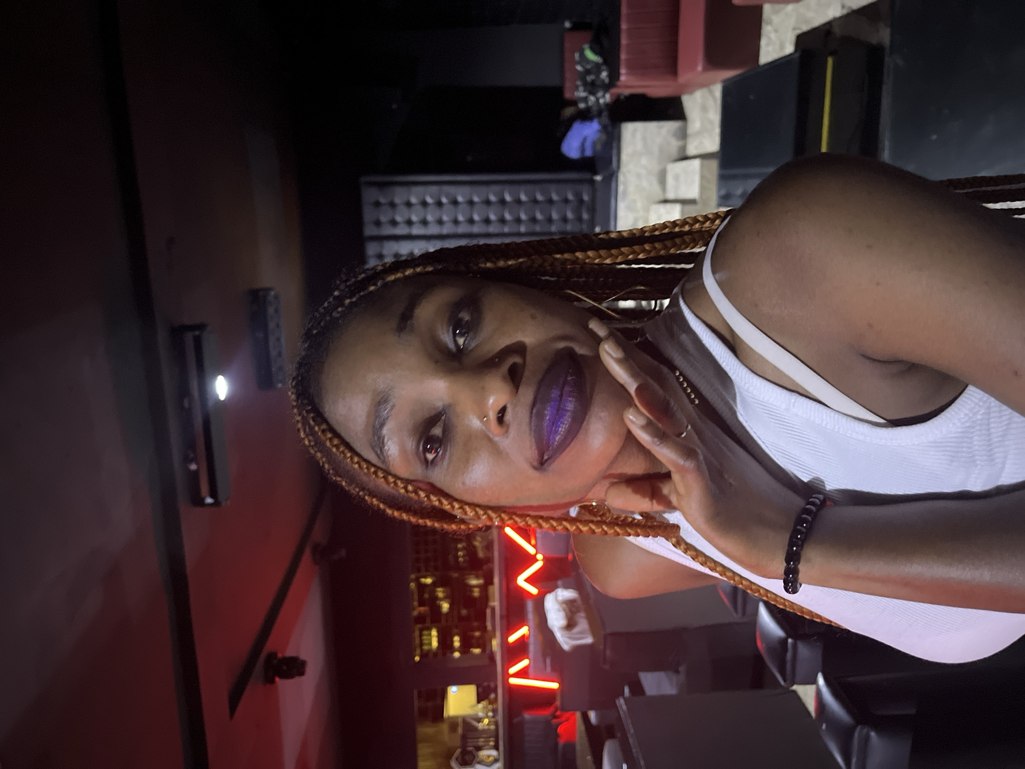 The image shows a medium shot of a woman with braided hair and dark purple lipstick. She is wearing a white tank top and a dark beaded bracelet. Her hand is resting on her chin, and she is looking towards the camera. The background appears to be a dimly lit indoor space, possibly a bar or lounge, with red neon lights and seating visible. The overall ambiance is dark and atmospheric.