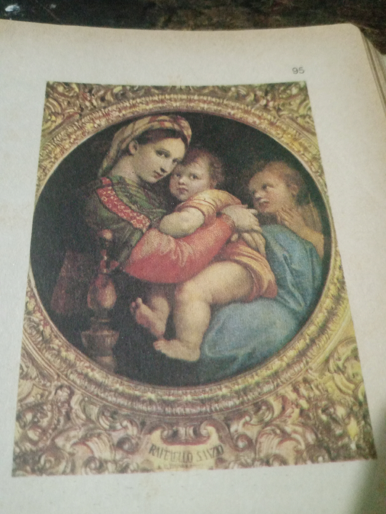 The image shows a close-up of a painting, likely a reproduction, set within an ornate oval frame. The painting depicts three figures: a woman holding a baby, with another young child leaning towards them. The woman on the left has dark hair and is wearing a green and red garment. She is looking down at the baby she cradles. The baby is swaddled in an orange cloth and has chubby cheeks. To the right, another child, possibly a girl with blonde hair, is looking at the baby and reaching out towards it. The figures are rendered in a soft, classical style. Below the painting, within the frame, is text that appears to read "RAFAELLO SANZIO". At the top right of the page, the number "95" is visible. The image is not a photograph taken in Santa Ana, Venezuela, but rather a reproduction of a painting.