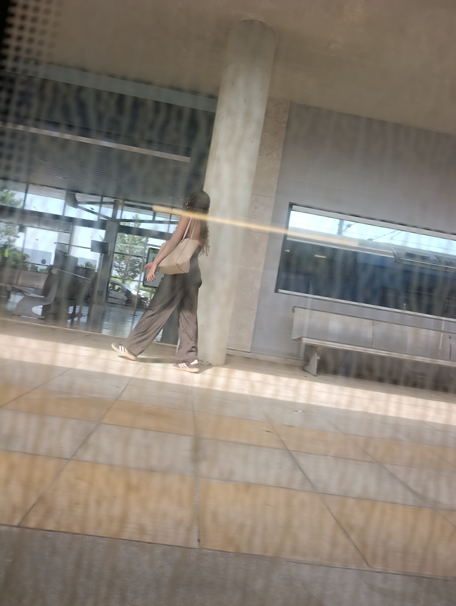 A woman with curly brown hair is walking from left to right in a spacious, modern-looking area with large windows. She is wearing a dark-colored jumpsuit and white sneakers with black stripes. She is carrying a light-colored tote bag over her right shoulder. Her hands are clasped in front of her. The scene appears to be an indoor waiting area or concourse, possibly a train station or airport terminal, due to the presence of benches and large glass windows. The floor is tiled with alternating light brown and cream-colored squares. Sunlight is streaming in through the windows, casting long shadows. The overall mood is calm and ordinary, with no discernible emotional expression from the woman. The time of day appears to be daytime, likely late morning or afternoon, given the bright sunlight. The weather outside seems clear. There is no visible text in the image.