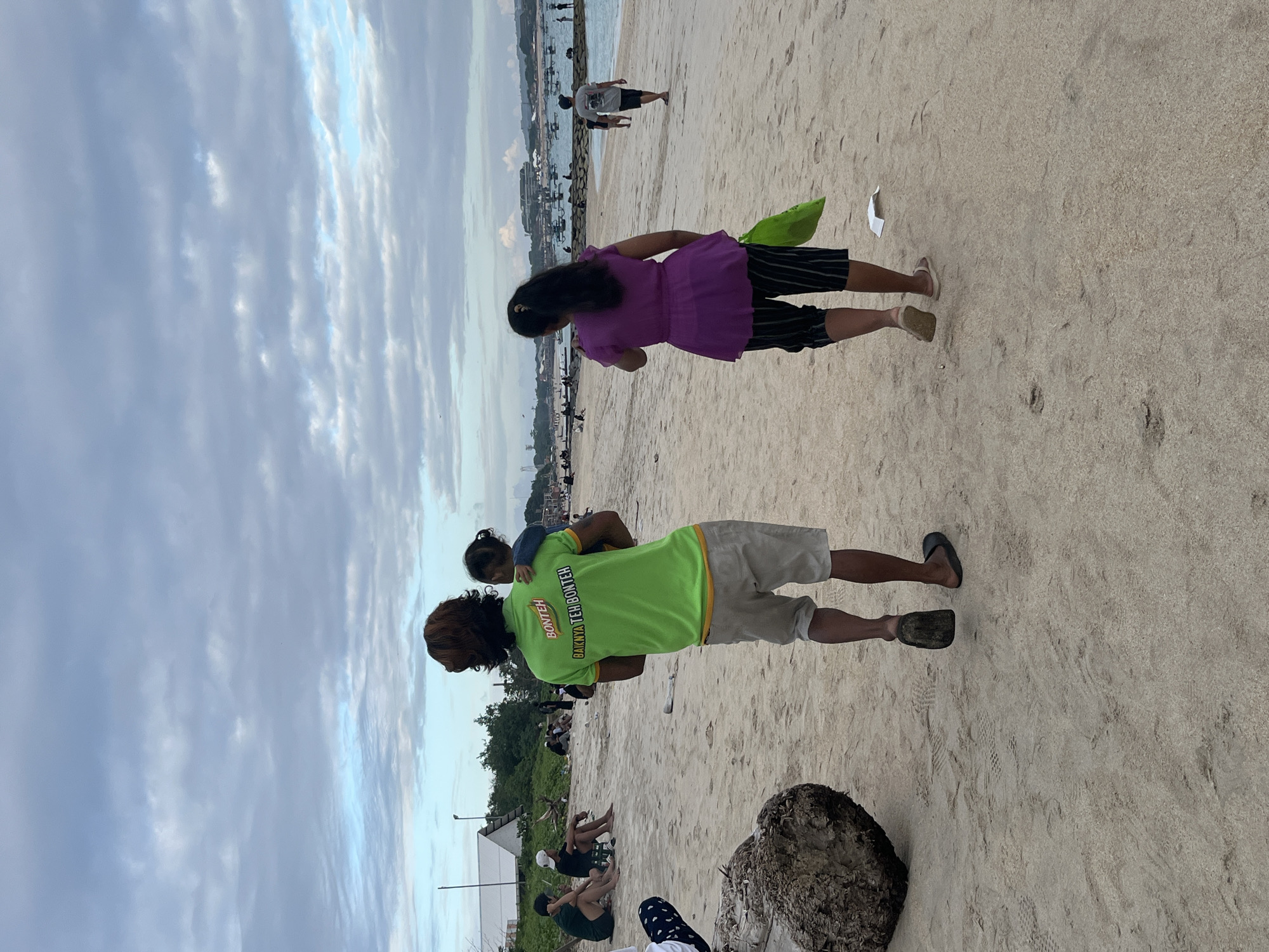 In Kuta, Indonesia, a family strolls along a sandy beach under a cloudy sky. A man in a bright green shirt, carrying a child on his hip, walks towards the ocean. The shirt bears the text "BONTEH BAIKNYA TEH BONTEH". Beside him, a woman in a purple top and striped shorts walks with a green bag. In the background, other beachgoers are visible, as are distant structures and boats. The environment is a typical beach scene with sand, water, and a hazy sky.