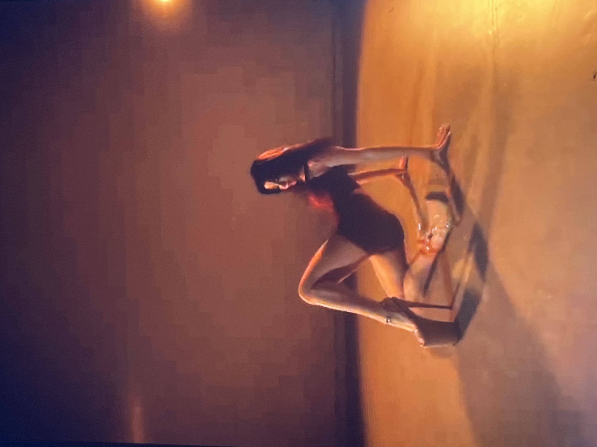A woman with long, dark hair is on the floor in a bent-over position, with her right leg extended and her left leg bent underneath her. She is wearing a dark, form-fitting outfit and very high platform heels. Her hands are on the floor in front of her, supporting her upper body. The environment appears to be an indoor space with a plain, light-colored wall in the background and a reflective, orange-toned floor. There is a bright light source in the upper right portion of the image, casting a warm glow. There is no indication of the location being in the city of Zoriane, Ukraine.