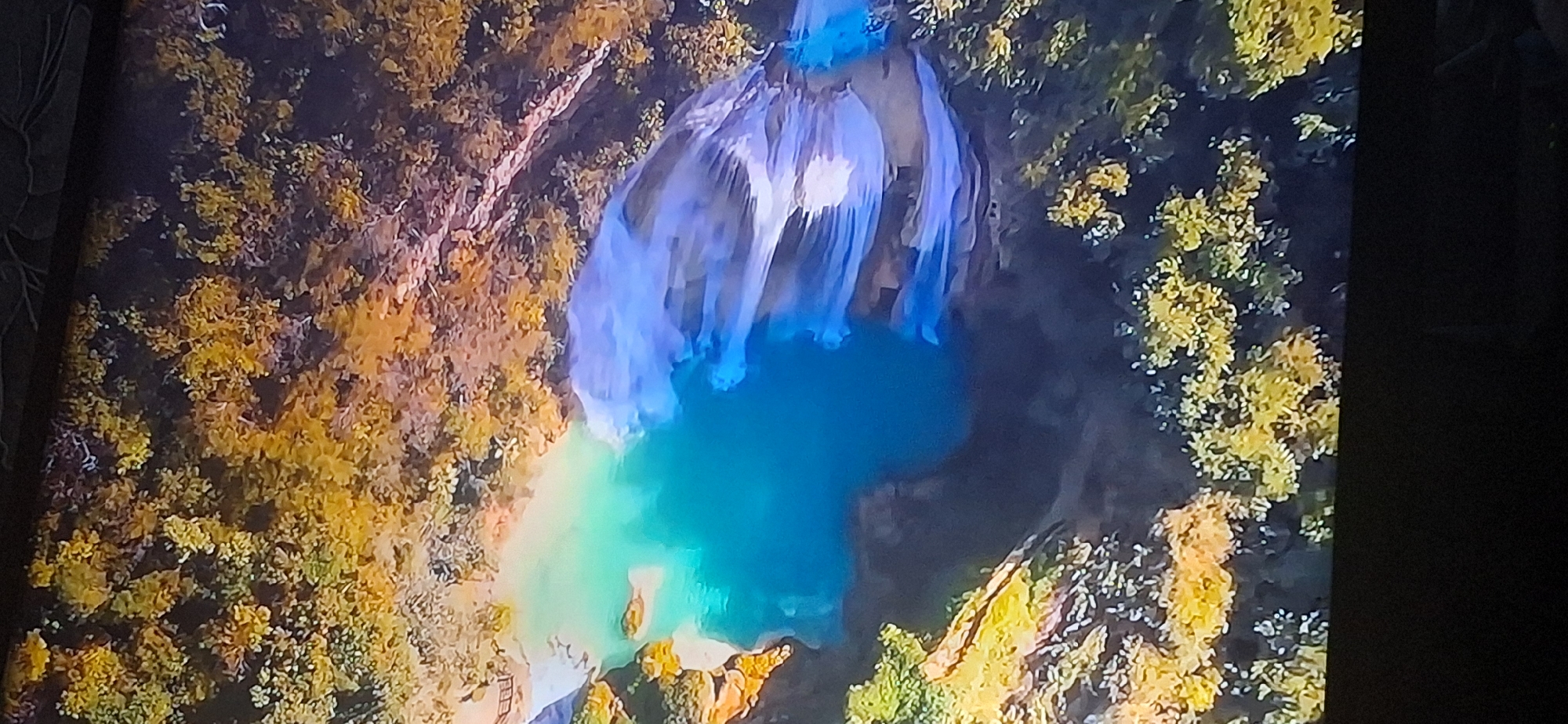 The image is an aerial view of a natural landscape featuring a vibrant turquoise pool of water. Surrounding the water is dense foliage with trees displaying a mix of green and golden-yellow leaves, suggesting an autumn or late summer season. The water appears to be a significant feature, possibly a lake or a deep part of a river, with a darker, possibly deeper, section transitioning into lighter, shallower areas near the edge. The overall scene conveys a sense of natural beauty and tranquility. There is no indication of the city of Bila Tserkva, Ukraine, in this image.
