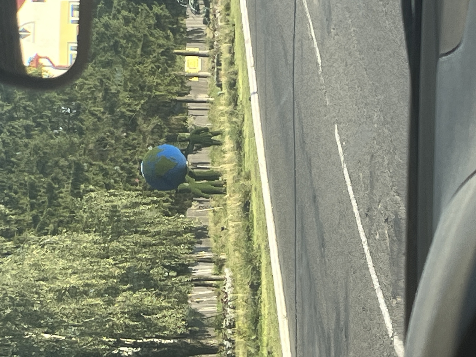 A decorative display features three green shrub figures holding up a large blue and green globe, representing Earth. The figures are arranged in a line, with two in the front and one slightly behind and to the right. Behind them, a line of tall trees forms a backdrop, with a wooden fence visible in front of some of the trees. The scene is set on a grassy area next to a road, with a white line marking the edge of the pavement. In the upper right corner, a reflection in a car mirror shows a yellow building with red trim. The location is the city of Lahojsk, Belarus.