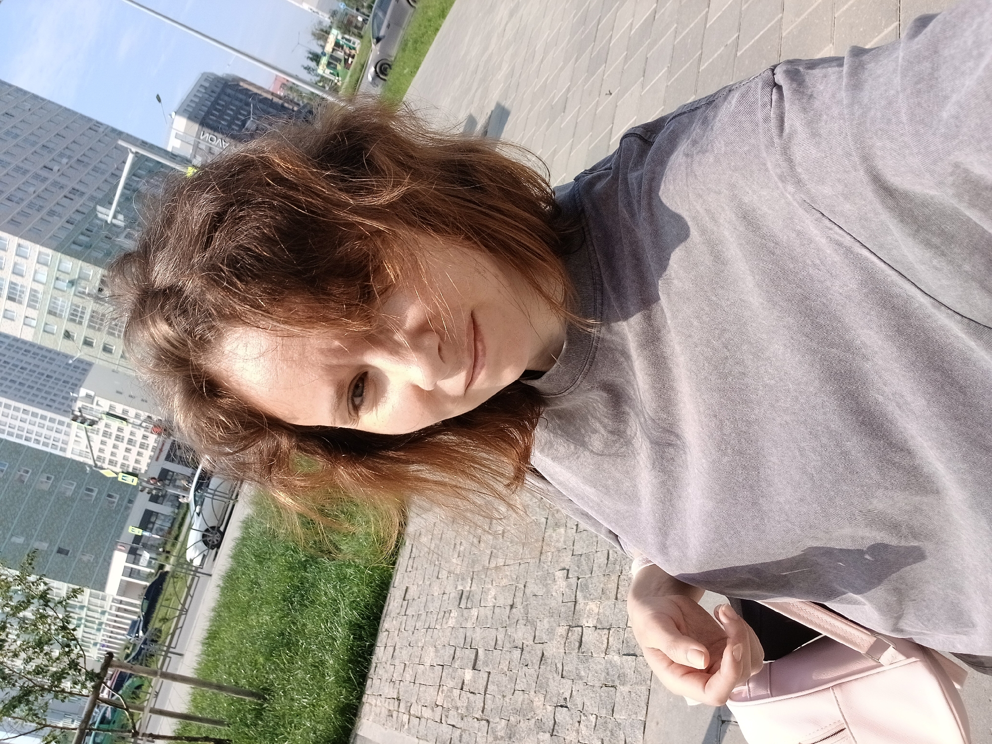 A young woman with short, reddish-brown hair is taking a selfie outdoors on a sunny day in a modern urban environment. She is wearing a grey t-shirt. A light pink backpack is visible on her left side. The background consists of tall, modern buildings, a street with cars, a grassy area, and a sidewalk paved with rectangular stones. The scene appears to be in Yekaterinburg, Russia, potentially near the "Europe-Asia" business center or a similar residential and commercial development.