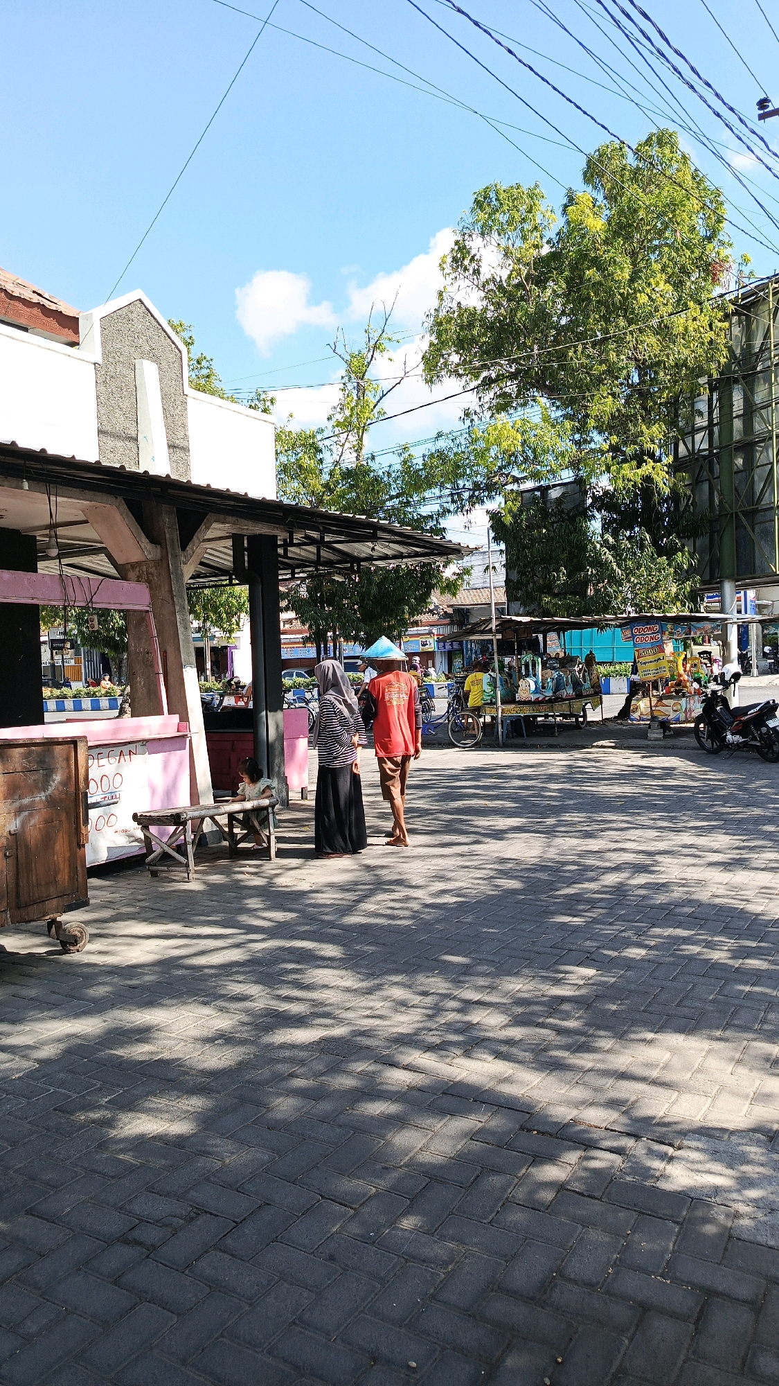 This image depicts an outdoor scene in the city of Nologaten, Indonesia, likely a marketplace or street with vendors. Two individuals, one wearing a red shirt and the other a hijab with a striped top and black skirt, are walking away from the camera. The person in the red shirt has a conical blue hat. In the foreground, a small child is seated on a wooden bench in front of a pink food stall. Other food stalls and parked motorcycles are visible in the background, along with trees and overhead power lines. The ground is paved with interlocking bricks, and dappled sunlight casts shadows across the scene.