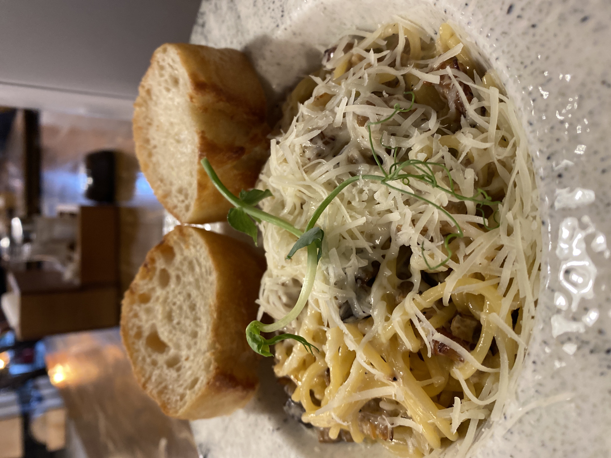 The image shows a plate of pasta with two slices of bread, likely in a restaurant setting. The pasta is topped with shredded cheese and a green garnish. The slices of bread are positioned next to the pasta. The plate has a speckled pattern. In the blurred background, there are objects suggesting a restaurant interior, including wooden blocks, possibly part of the decor, and reflective surfaces. The location is not discernible as Yekaterinburg, Russia.