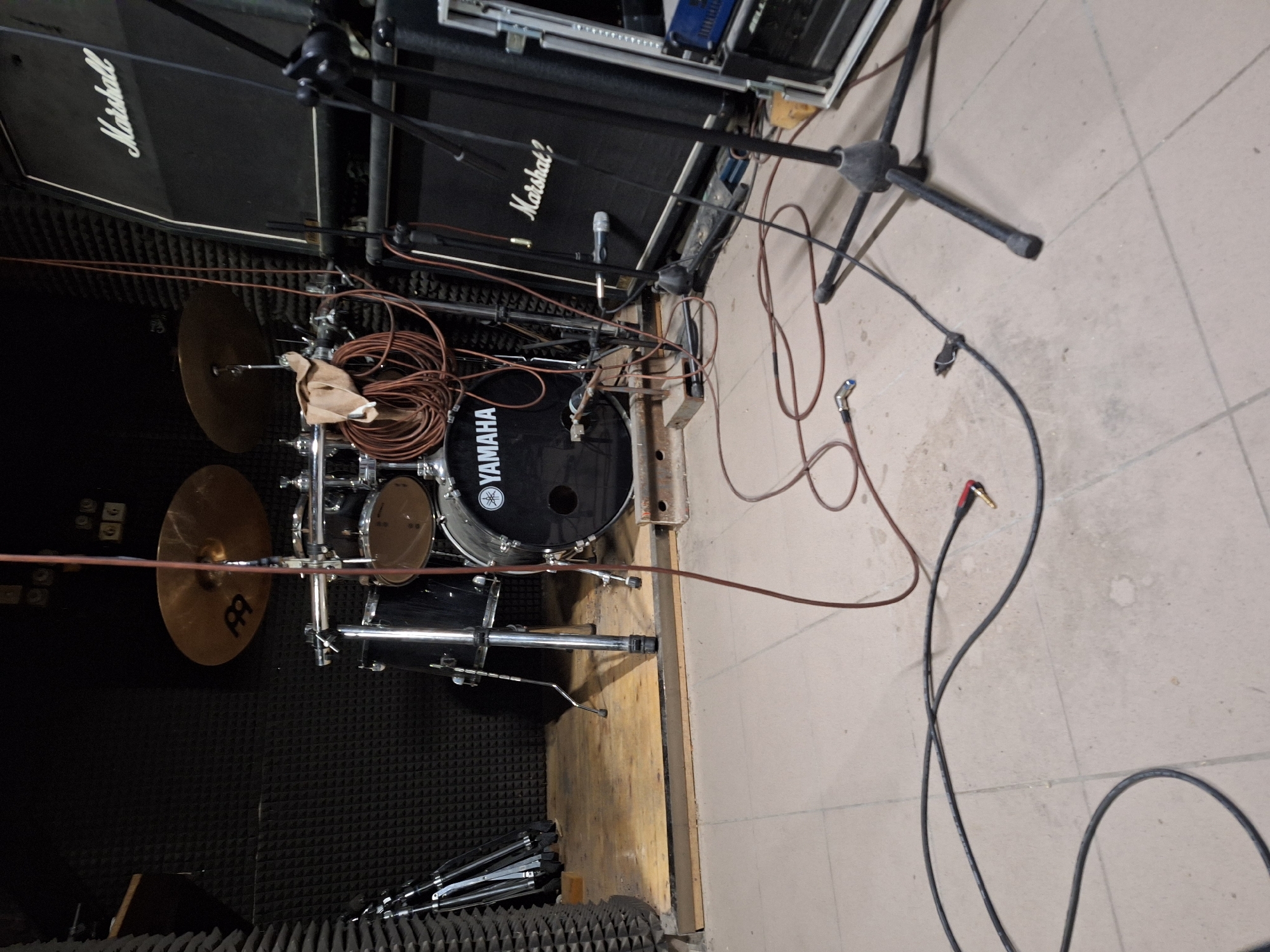 The image shows a Yamaha drum kit in a room with soundproofing foam on the walls. The drum kit includes cymbals, toms, a snare drum, and a bass drum. Two Marshall amplifiers are positioned behind the drum kit. Several microphones and stands are visible, along with various cables. The floor is tiled. The image was taken in Panagyurishte, Bulgaria.