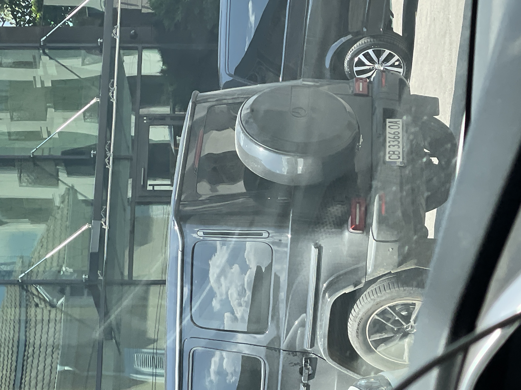 The image shows the rear view of a dark-colored Mercedes-Benz G-Class SUV parked on a paved surface, likely a street or parking lot. The SUV has a spare tire mounted on its rear door and a license plate that reads "CB 3366 OA". To the right of the G-Class, the rear of a black van is partially visible. In the background, a modern building with large glass windows and a tiled roof is reflected in the SUV's windows. The sky with scattered clouds is also reflected. The scene appears to be set in an urban environment, consistent with the city of Sofia, Bulgaria, known for its mix of modern architecture and historical buildings. The lighting suggests it is daytime, possibly late morning or early afternoon, given the clear sky and sharp reflections. There are no people clearly visible in the image, though faint reflections within the SUV's windows might suggest occupants. The overall impression is a snapshot of urban life with vehicles and architecture.