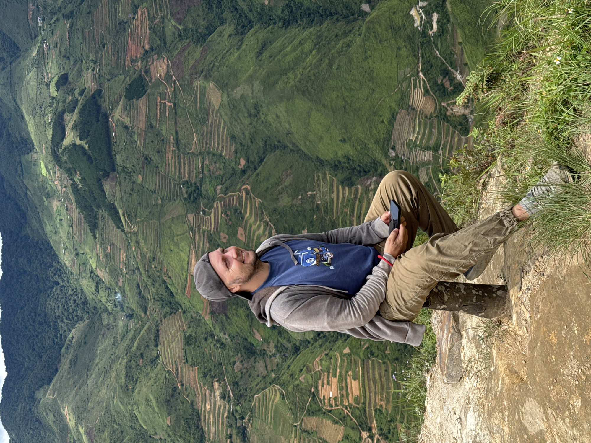 A man is sitting on a rustic wooden stool on a rocky outcrop overlooking a valley in Ta Xua, Vietnam. He is wearing a grey baseball cap, a blue t-shirt with a graphic print, a grey zip-up hoodie, and khaki pants. He is holding a smartphone in his right hand and looking to his right with a smile. The background is dominated by lush green terraced hillsides, characteristic of the region's rice cultivation. The sky is overcast, creating a soft light. The man appears relaxed and contemplative, enjoying the scenic vista.
