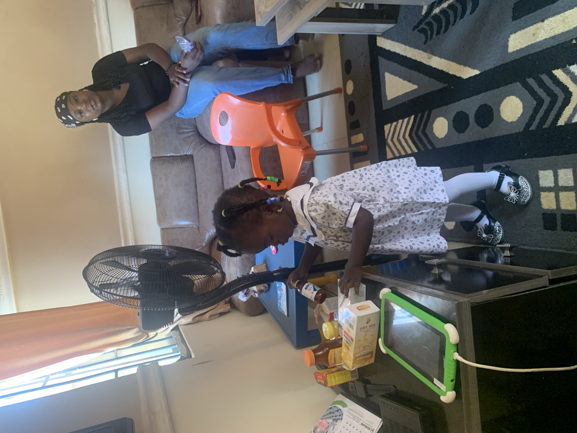 The image shows a young Black child wearing a white dress with a black and white pattern and black shoes with butterfly embellishments. The child has braided hair with beads. The child is on their knees on a floor with a rug featuring a geometric pattern. They are reaching for items on a black coffee table. On the table are several boxes, including one labeled "BIOMED FOR KIDS" with orange text and a cartoon illustration. There are also some bottles with orange and yellow labels. A green tablet with white bumpers is also on the table. To the left of the child, a black oscillating fan is visible.
In the background, a woman in a black t-shirt and a black and white headscarf is seated on a brown leather couch. She is wearing blue jeans and has her arms around someone, holding a clear plastic bag. An orange plastic child's chair is next to the couch. The scene appears to be indoors, likely a living room, given the furniture and rug. The lighting suggests it could be daytime. The overall mood seems casual and domestic.