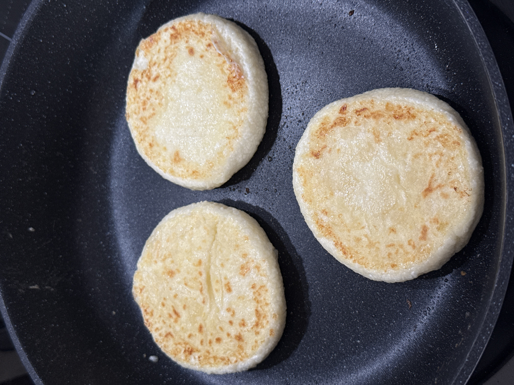 Three round, pale, flatbread-like items are being cooked in a black non-stick frying pan. The items have light golden-brown spots scattered across their surfaces, indicating they are browning. The pan is set on a stovetop, visible at the top edge of the image. The environment is likely a kitchen in a residential setting. There are no people visible in the image. The image is cropped to focus on the food and the pan.