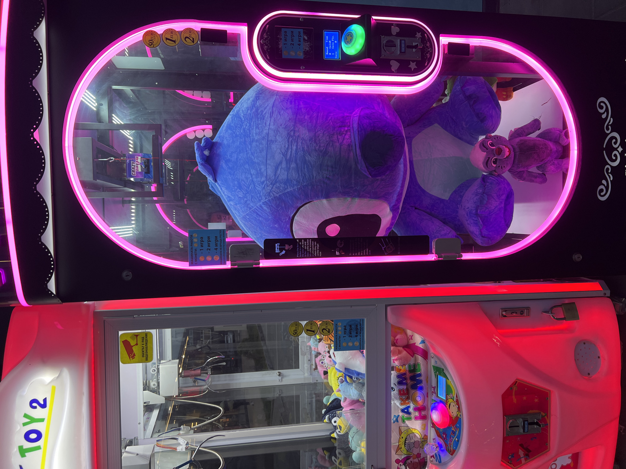 The image captures two brightly lit claw machines in an indoor setting, likely an arcade or entertainment venue in Plovdiv, Bulgaria.
The prominent upper machine features vibrant pink neon lighting outlining its large viewing window. Inside, a substantial blue/purple plush toy, closely resembling Disney's Stitch, is displayed on its back. To its right, a smaller, dark purple, monkey-like plush toy is also visible. The machine's control panel is illuminated with pink and blue lights, showing coin slots for 50 euro cents, 1 euro, and 2 euros. A digital display reads "Round," "Time," "Coin," and "1 Coin Pls." An information panel in Bulgarian states: "МАШИНАТА РАБОТИ С МОНЕТИ: 1 игра, 2 игри, 4 игри" (The machine works with coins: 1 game, 2 games, 4 games), and "МАШИНАТА НЕ ВРЪЩА РЕСТО" (The machine does not return change). A metallic claw mechanism hangs above the plush toys.
Below, a smaller claw machine with white and red trim is also internally lit. It contains numerous smaller plush toys of various animals, including pigs and cats. "TOY 2" is visible on its side, and a colorful banner inside reads "TAKE ME HOME." This machine also features coin slots for 50 euro cents, 1 euro, and 2 euros, with identical Bulgarian pricing and change information. A yellow sticker on the glass warns, "ОБЕКТЪТ Е ПОД ВИДЕОНАБЛЮДЕНИЕ" (The object is under video surveillance). The machines are currently empty of players, their inviting glow awaiting interaction.