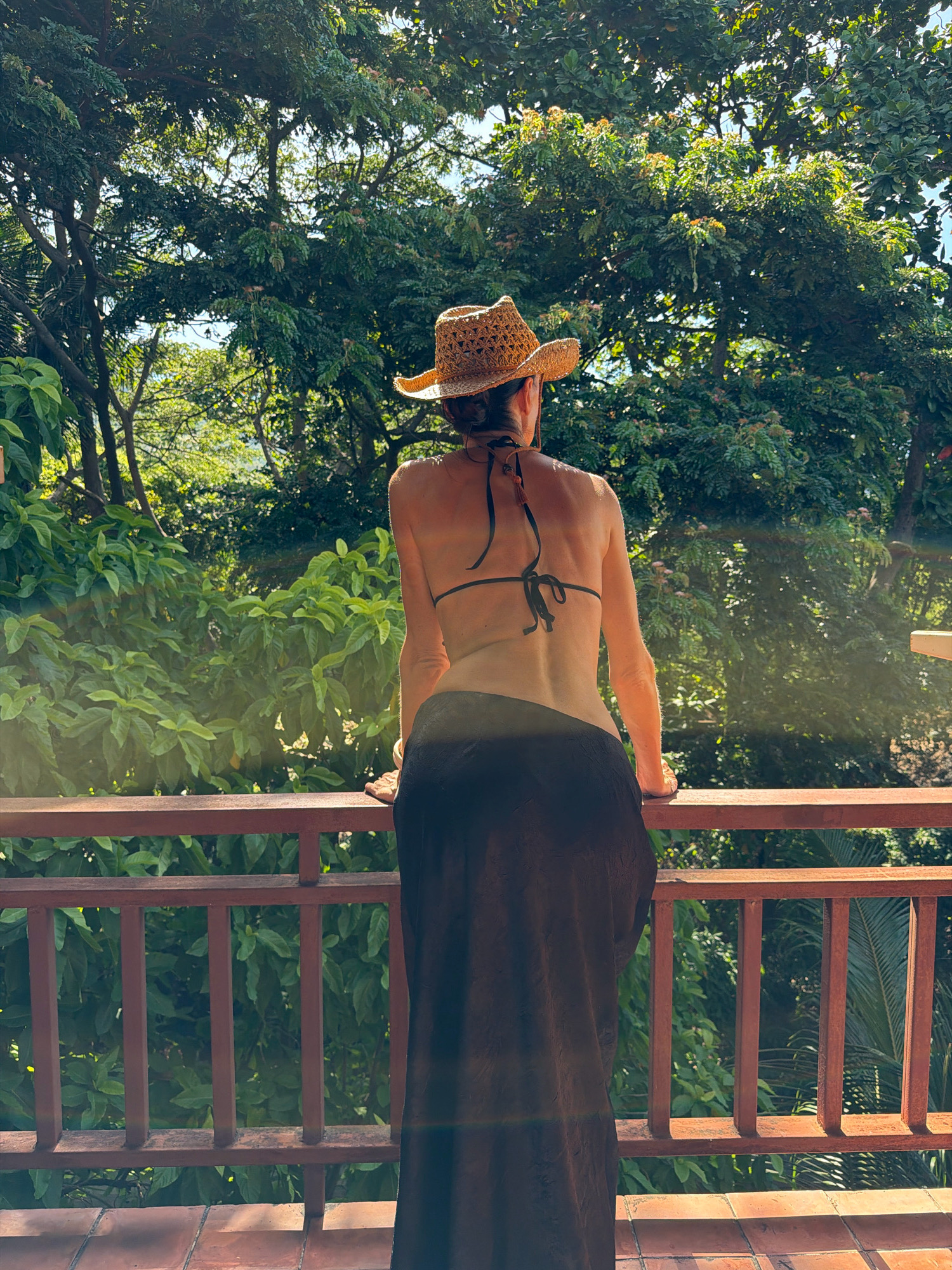 A woman wearing a straw hat and a black bikini top with a black skirt stands on a wooden balcony, leaning on the railing. She is viewed from behind, with her back to the camera. Lush green trees and foliage surround the balcony. The image appears to be taken on a sunny day, with a lens flare visible in the foreground. The location is the city of Ko Samui District, Thailand.