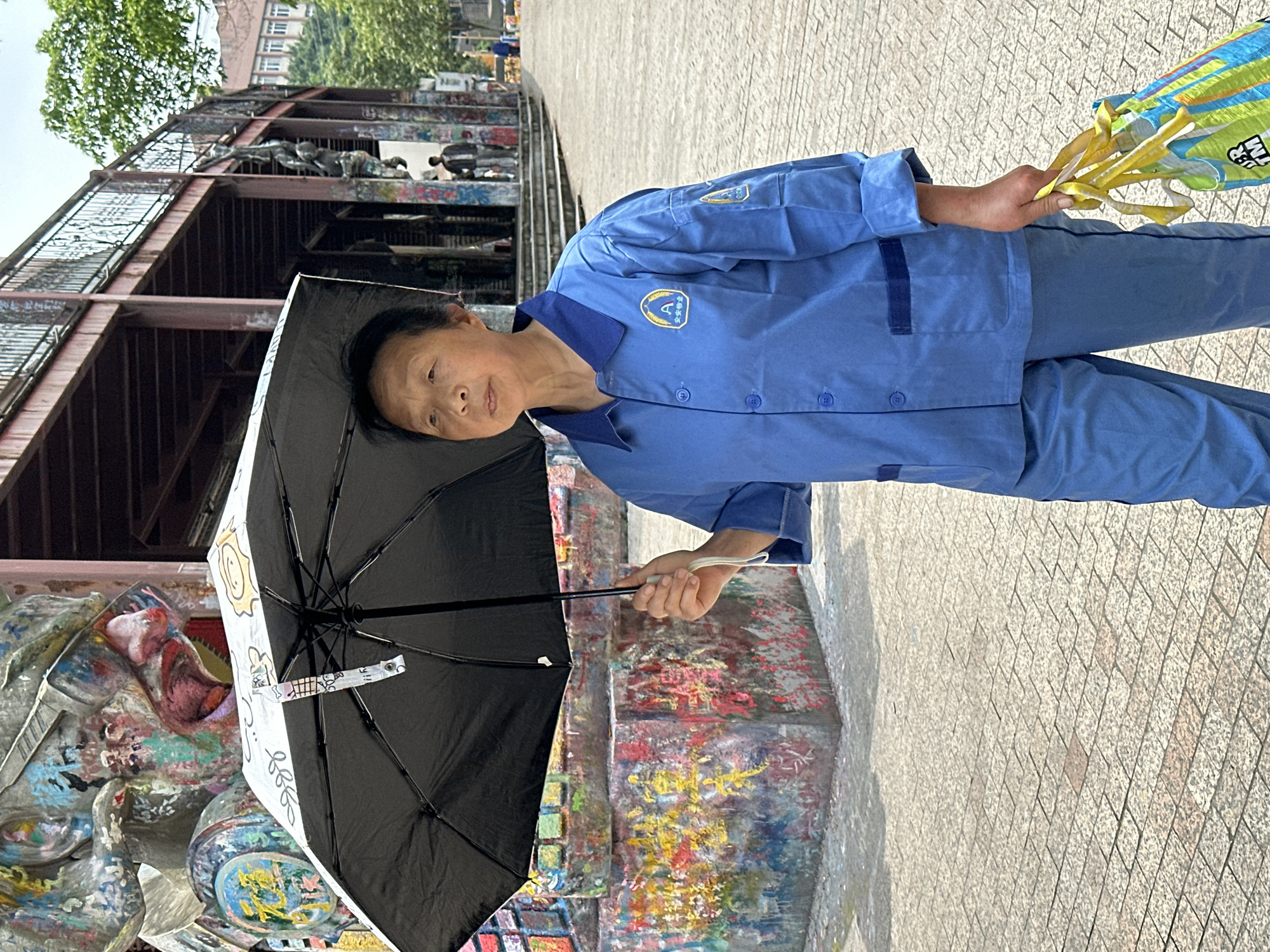 A middle-aged woman stands on a tiled walkway in Jiulongpo District, China, holding a black umbrella decorated with white cartoon images and a bundled item wrapped in a blue, green, and yellow patterned plastic bag. She is wearing a blue uniform shirt and pants with darker blue trim and a patch with a shield symbol on the upper left chest. Behind her is a painted, graffiti-covered structure and a metal framework supporting a walkway or bridge with figures on it. The weather appears bright and sunny.