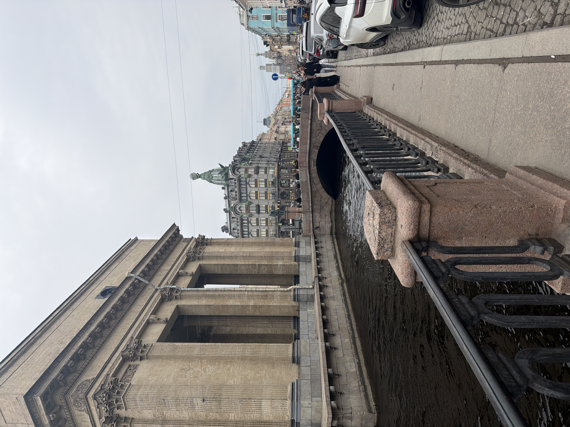 The image depicts an urban scene along the Griboyedov Canal in Saint Petersburg, Russia, under an overcast sky.
In the foreground, a dark metal railing with decorative elements is affixed to a reddish-brown granite balustrade. The Griboyedov Canal runs behind this barrier, displaying dark, rippled water. To the left, the monumental facade of the Kazan Cathedral, constructed from light beige stone, features a series of prominent Corinthian columns and a detailed entablature. The base of the cathedral is grey stone, and a dark, rectangular opening is visible on its upper wall.
On the right side of the image, a concrete sidewalk runs parallel to the canal, leading to a cobblestone street where several cars are parked, including a white SUV and a grey sedan. Pedestrians are scattered along the sidewalk and a stone arch bridge, identified as the Kazan Bridge, which spans the canal directly ahead. Two individuals are prominent on the right sidewalk near the railing: one wears a long dark coat, and the other a lighter jacket and jeans.
Beyond the Kazan Bridge, the Singer House (Dom Knigi) is a distinctive landmark with its ornate grey Art Nouveau architecture and a prominent green-domed tower topped with a spherical structure. Further down the street, a light blue bus or tram is in motion amidst other vehicles and a collection of multi-colored buildings. In the far distance, the partially obscured onion domes of the Church of the Savior on Spilled Blood are visible. Overhead, utility lines traverse the sky.