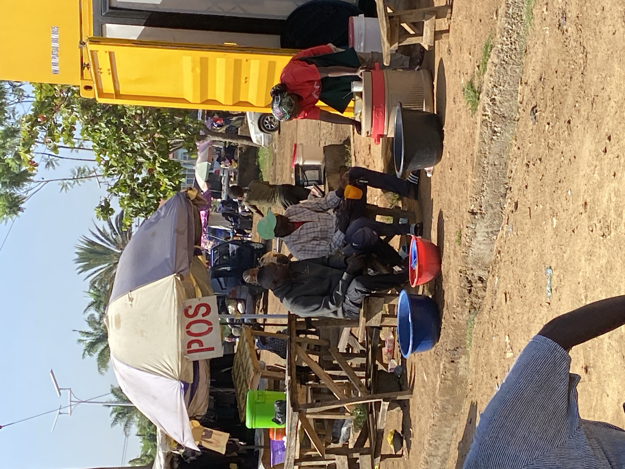 The image shows a bustling scene in Bukuru, Nigeria. Several people are gathered around a yellow structure resembling a shipping container, potentially repurposed for commercial use. One woman in a red shirt is actively working with a large container, possibly dispensing liquids, while several men are seated on wooden chairs and benches nearby. A white umbrella with the red letters "POS" stands out, suggesting a point-of-sale service is available. Plastic buckets of various colors (blue, red, green) are scattered around. The ground is dry and dusty. A person in a blue-striped shirt is partially visible in the foreground. Trees are visible in the background under a sunny sky.