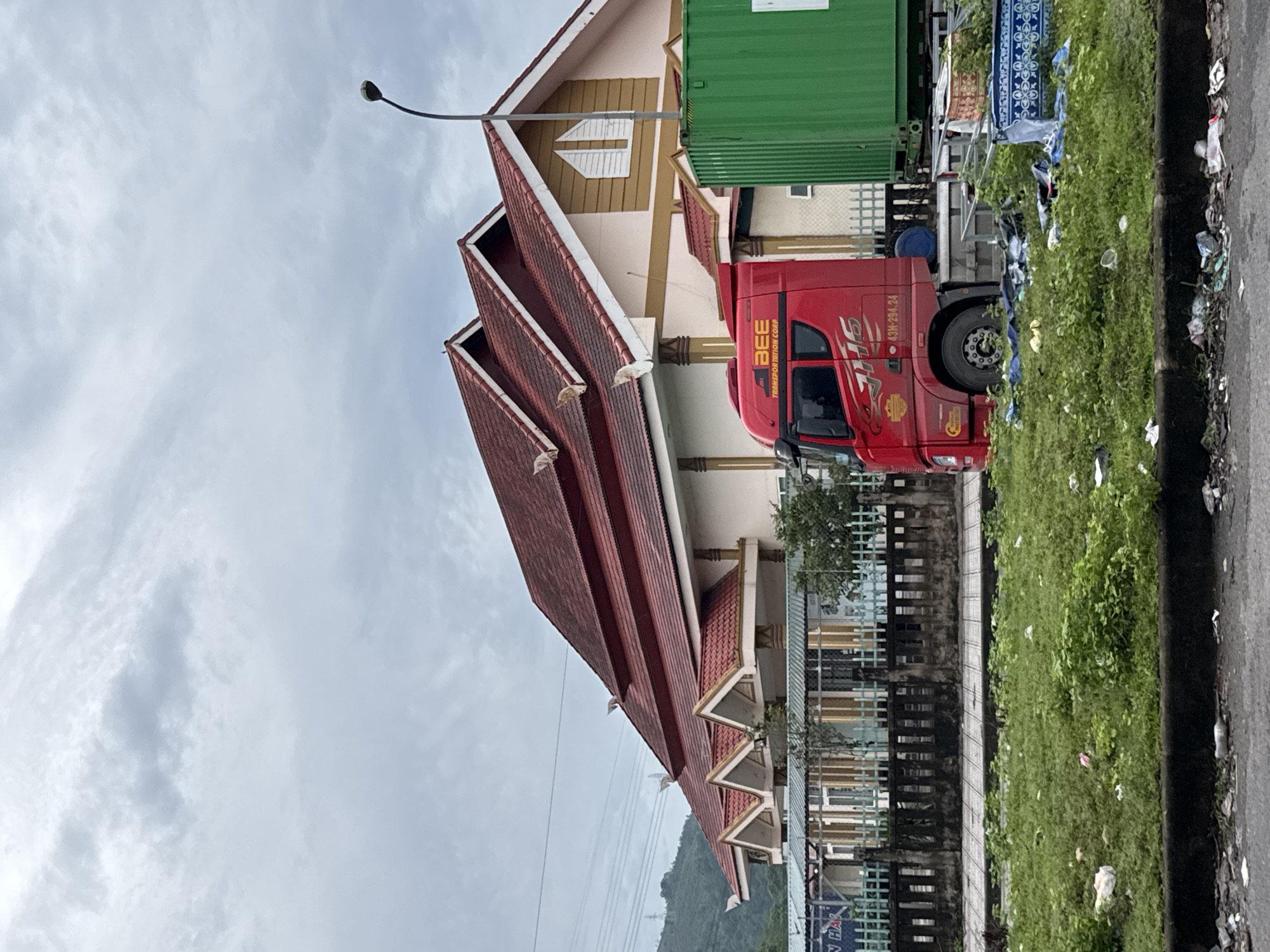 The image depicts a red truck with "BEE TRANSPORTATION CORP" printed on its side, parked near a building with a red, multi-tiered roof. A green shipping container is visible next to the truck. The scene is located in Lao Bảo Commune, Quảng Trị Province, Vietnam. The ground is covered in green grass with scattered litter, including plastic bags and bottles. A paved walkway is present behind the grass, and a fence separates the walkway from the building. The sky is overcast with clouds. The building appears to be a cultural or administrative center, possibly a temple or a pagoda, with distinctive architectural features. The overall scene suggests a commercial area adjacent to a significant structure, with evidence of urban decay due to the presence of litter.