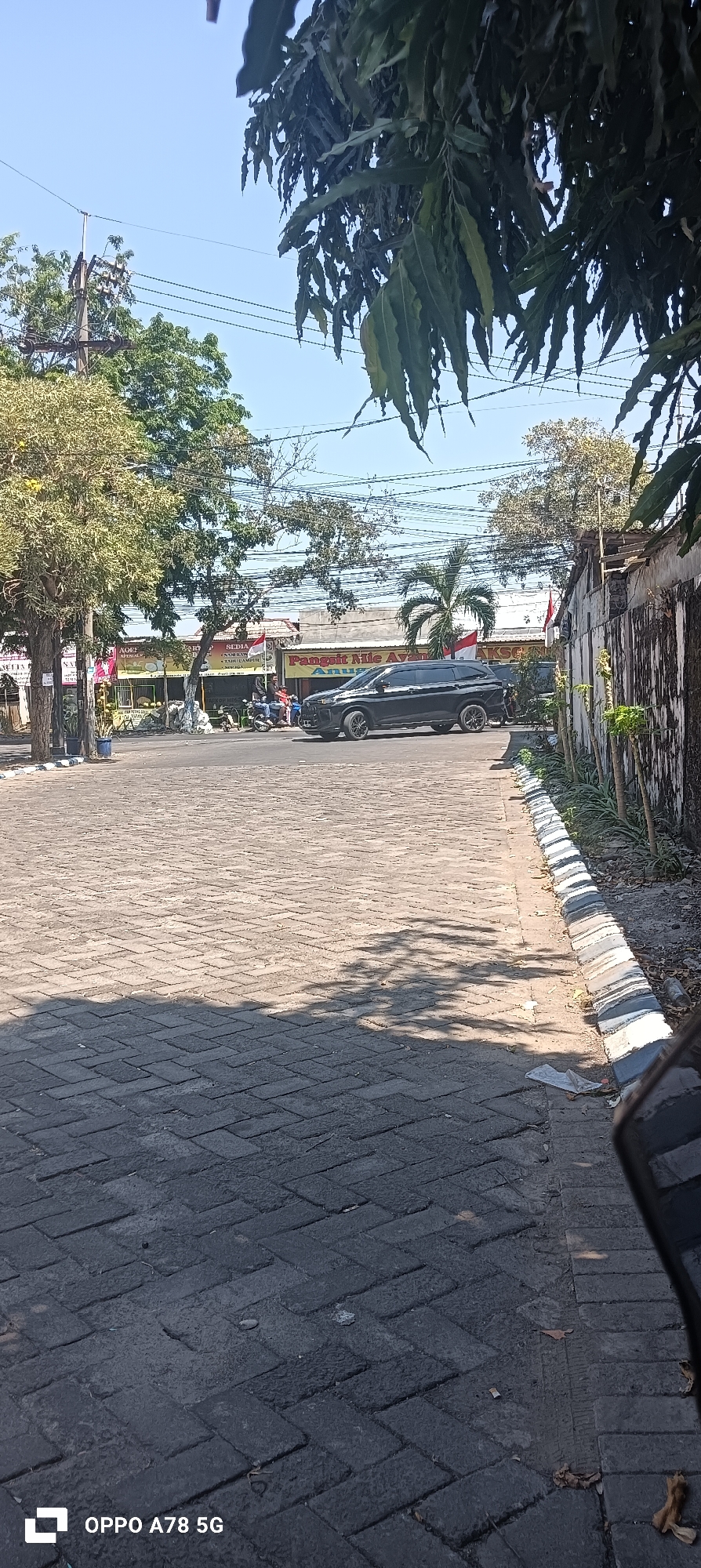 The image depicts a street scene in Kwangsan, Indonesia, on a clear, sunny day. The foreground is a paved area of dark grey brick pavers, with a distinct shadow cast by unseen foliage from the upper right. To the left, a narrow strip of lighter brick pavers borders a raised curb with alternating grey and white painted sections. Beyond this, a paved street runs horizontally across the frame.
In the middle ground, a dark colored SUV is parked or moving on the street. Behind the SUV, a row of low-rise buildings lines the street, adorned with banners and signs, some featuring Indonesian flags. Several individuals are visible on motorcycles in front of the buildings, and trees with green foliage are interspersed along the street and behind the buildings. Palm trees are also present.
The background shows a clear blue sky, with power lines and utility poles visible. To the right of the frame, a textured wall, possibly made of concrete or brick, runs vertically along the edge of the property, with some sparse vegetation growing along its base. The overall impression is of a typical urban street in a tropical climate, with commercial establishments and infrastructure typical of Indonesia.