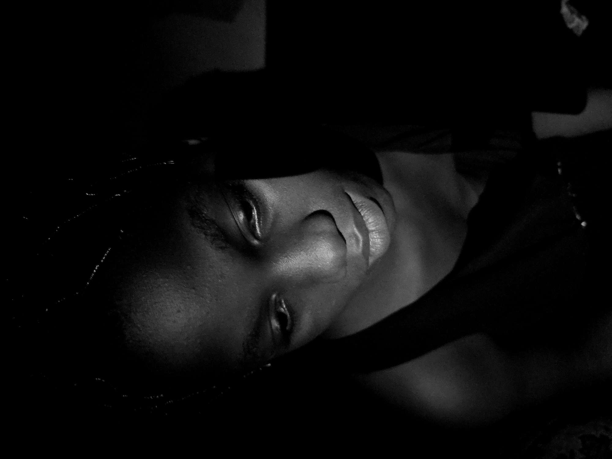The image is a black and white, close-up of a person lying down. The subject's face is the most prominent feature, with their eyes partially open and lips slightly parted. Light is selectively illuminating the face, creating high contrast with the surrounding shadows. Fine details such as hair braids are visible on the left side of the frame. The background is predominantly dark, obscuring details. The absence of contextual clues makes it impossible to determine the location in Asaba, Nigeria, or any other specific details about the environment.