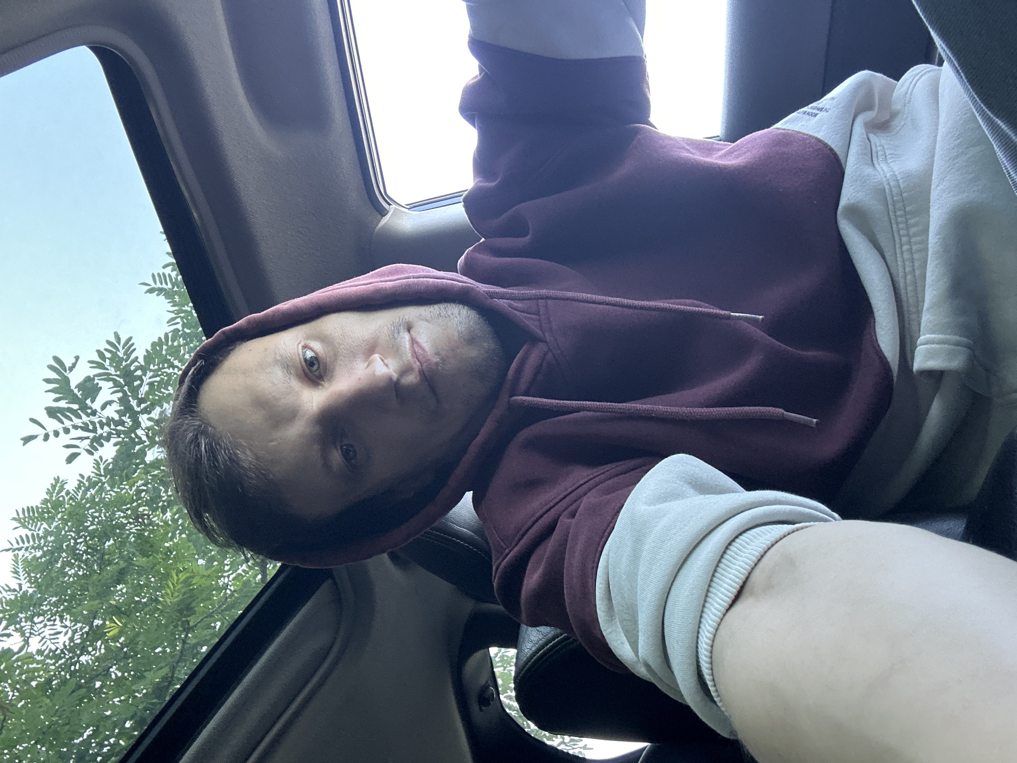 A man is taking a selfie inside a car with the sunroof open. He is wearing a maroon hoodie with light-colored sleeves. Green foliage and the sky are visible through the sunroof. The interior of the car, including dark leather seats, is also visible. The image is a close-up shot, focusing on the man's face and upper body. The scene appears to be outdoors, possibly in a rural or suburban area given the presence of trees. The man's facial expression is neutral. There is no specific information about the location in the city of Makiv, Ukraine.