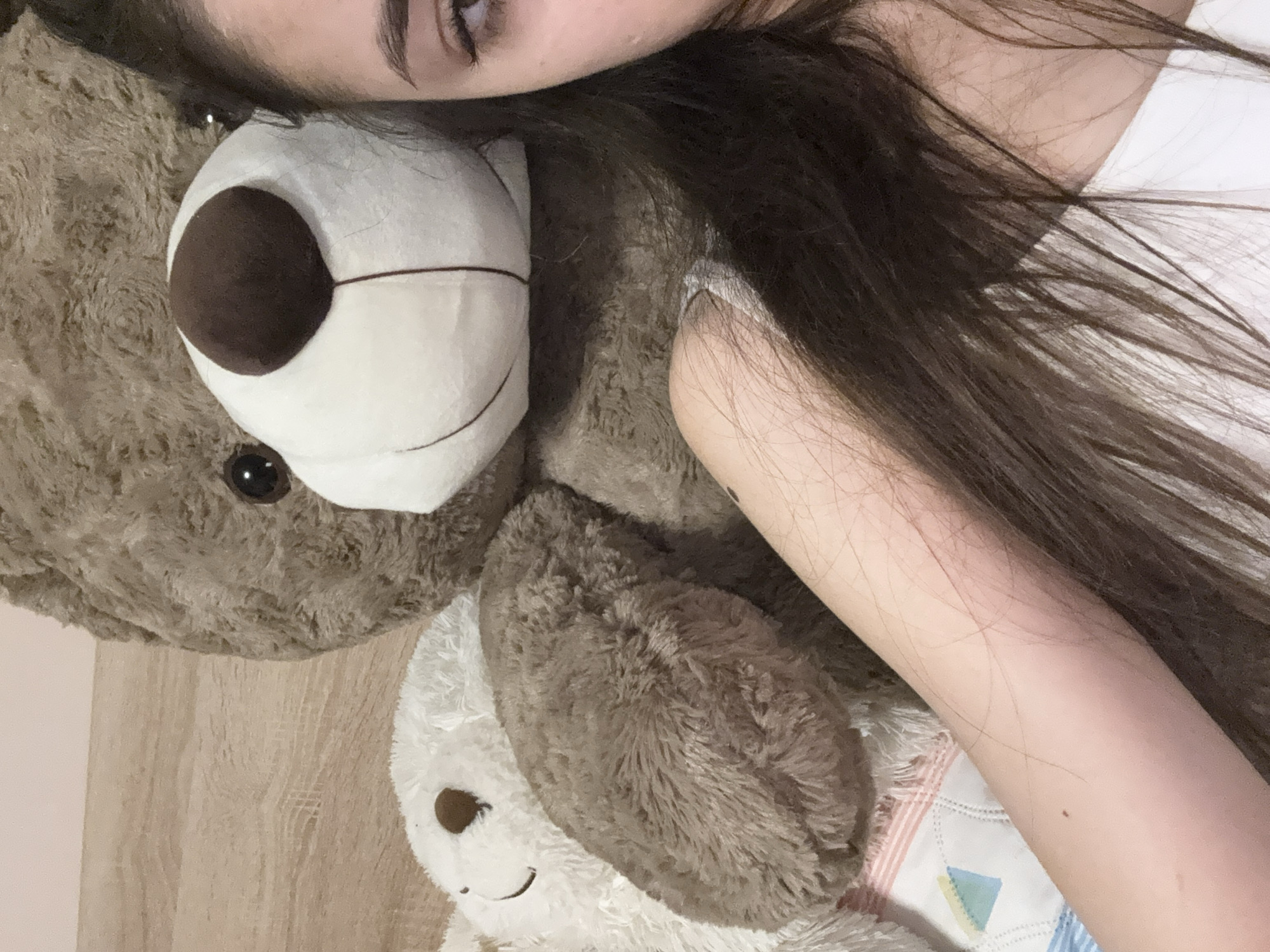 A young woman is lying down with a large brown teddy bear and a smaller white teddy bear. The woman is partially visible with dark hair, fair skin, and a visible eye. She is wearing a white top. The teddy bears are lying on top of what appears to be a bed or blanket with a pastel pattern. The background includes a wooden surface, likely a floor or wall. The image does not provide enough context to confirm the location in Iarebichna, Bulgaria.