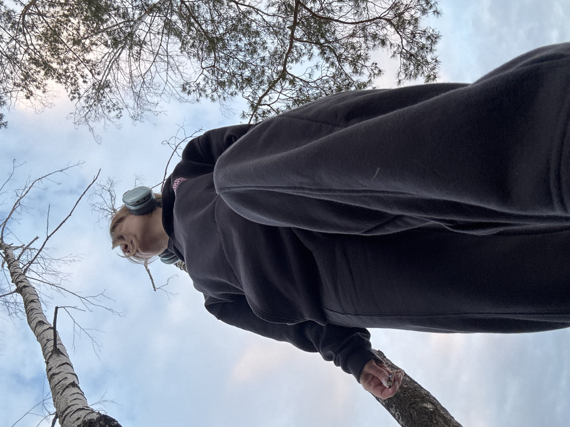 A low-angle outdoor photograph depicts an individual, appearing female with light-colored hair, standing within a wooded area in the city of Noviy, Russia. The person is dressed in a dark hooded sweatshirt and matching dark sweatpants, wearing light-colored over-ear headphones. Their head is tilted upwards, looking towards the sky. The individual's right hand is visible, gently grasping the trunk of a birch tree, with a small, light-colored ring present on a finger. The background consists of a partially cloudy blue sky and various tree branches, including a prominent birch tree with bare branches on the left and a pine tree with needles on the right.