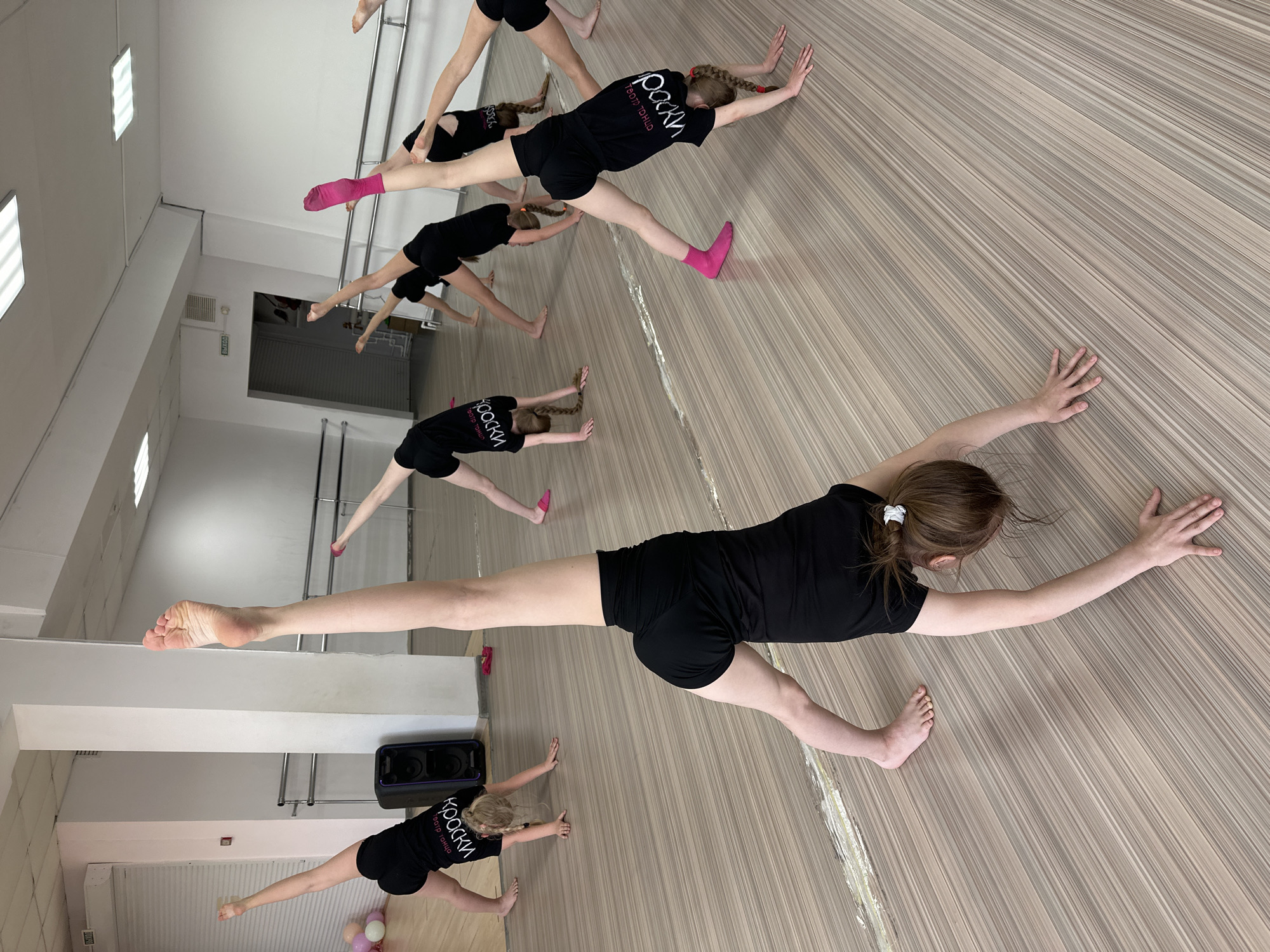 A group of young female dancers are stretching in a dance studio in Votkinsk, Russia. The studio has a wood-patterned floor and mirrors along one wall. The dancers are wearing black shirts and shorts, with some wearing pink socks. They are performing a stretching exercise, with their legs extended to the sides and arms reaching forward.