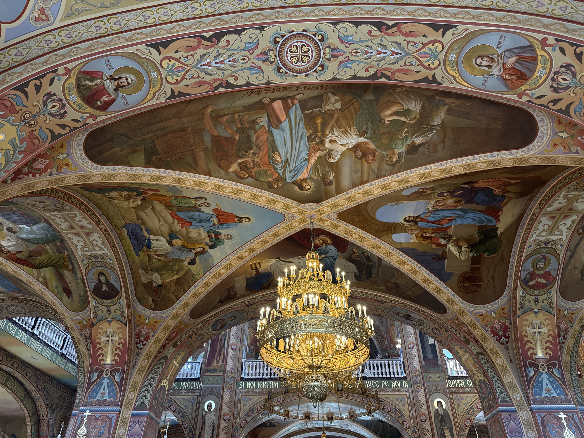The image shows the ornate interior of a church, focusing on the elaborately painted ceiling and a large, gilded chandelier. The ceiling is adorned with frescoes depicting religious scenes and figures, with intricate decorative borders in various colors and patterns. The frescoed sections are separated by arched architectural elements. The chandelier, hanging prominently in the center, is a multi-tiered, ornate gold fixture with numerous lit candles, casting a warm glow on the surroundings. Below the ceiling, balconies with white railings are visible, with people standing and observing. The architecture suggests a grand and traditional religious space, possibly an Orthodox church, given the style of the iconography in the frescoes. The overall impression is one of rich decoration and spiritual grandeur.