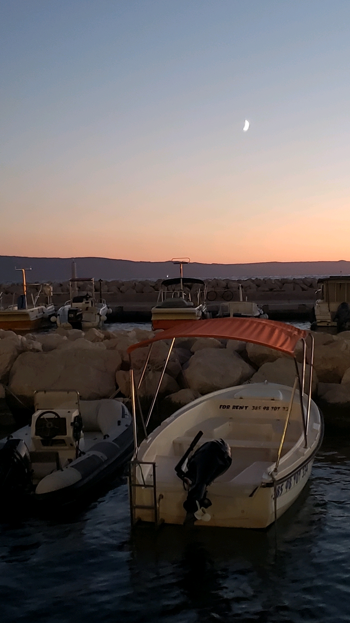 The image depicts a serene sunset scene at a harbor in Općina Tučepi, Croatia. The sky transitions from a light blue at the top to a soft orange and pink hue near the horizon, with a sliver of a crescent moon visible. The foreground is dominated by several boats docked among large, light-colored rocks.
In the immediate foreground, a white boat with an orange canopy is prominently featured. It has a black outboard motor and a metal ladder at the stern. The boat has the inscription "FOR RENT 385 98 707 334" on its side. To its left, an inflatable boat with gray and white detailing, also with a black outboard motor, is partially visible.
Behind these boats, several other vessels are docked, some with canopies. These appear to be smaller boats, possibly also for rent, typical of a tourist area. In the middle ground, a rocky breakwater separates the harbor from the sea. Beyond the breakwater, the calm sea reflects the colors of the sky. In the distance, a dark, low-lying landmass, likely hills or mountains, forms the horizon. A small lighthouse or tower can be seen on the breakwater. The overall atmosphere is peaceful and tranquil, characteristic of a coastal town at dusk.