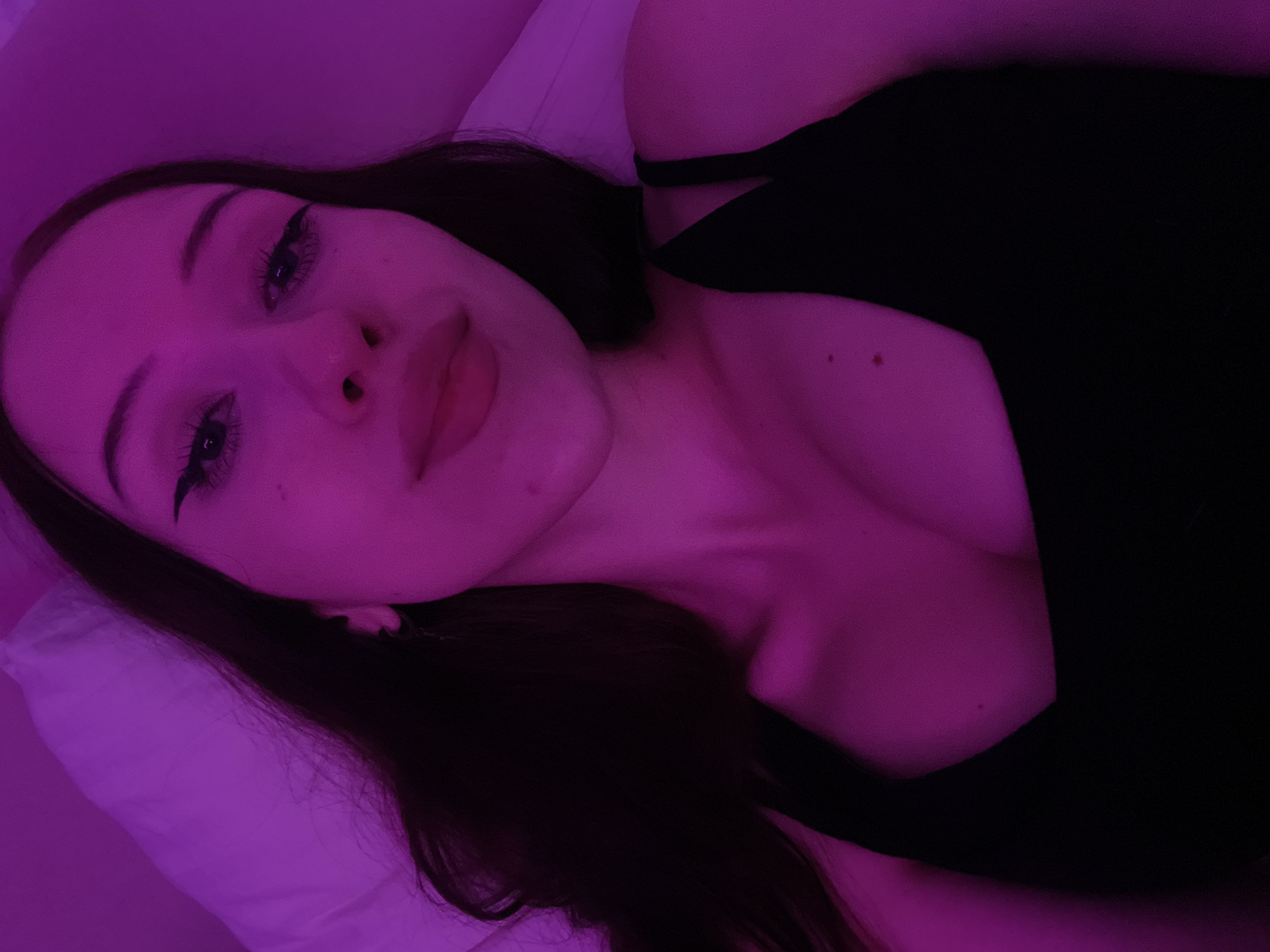 The image shows a close-up, upper-body selfie of a young woman with long, dark hair. She is lying down, with a white pillow visible behind her head. The scene is illuminated by a pinkish-purple light, likely from an artificial source such as a neon light or LED lamp. The woman is wearing a black tank top, and her makeup includes winged eyeliner and mascara. The lighting casts a strong magenta hue over the entire image, obscuring fine details and the background. There is no information in the image to suggest a location in Votkinsk, Russia.