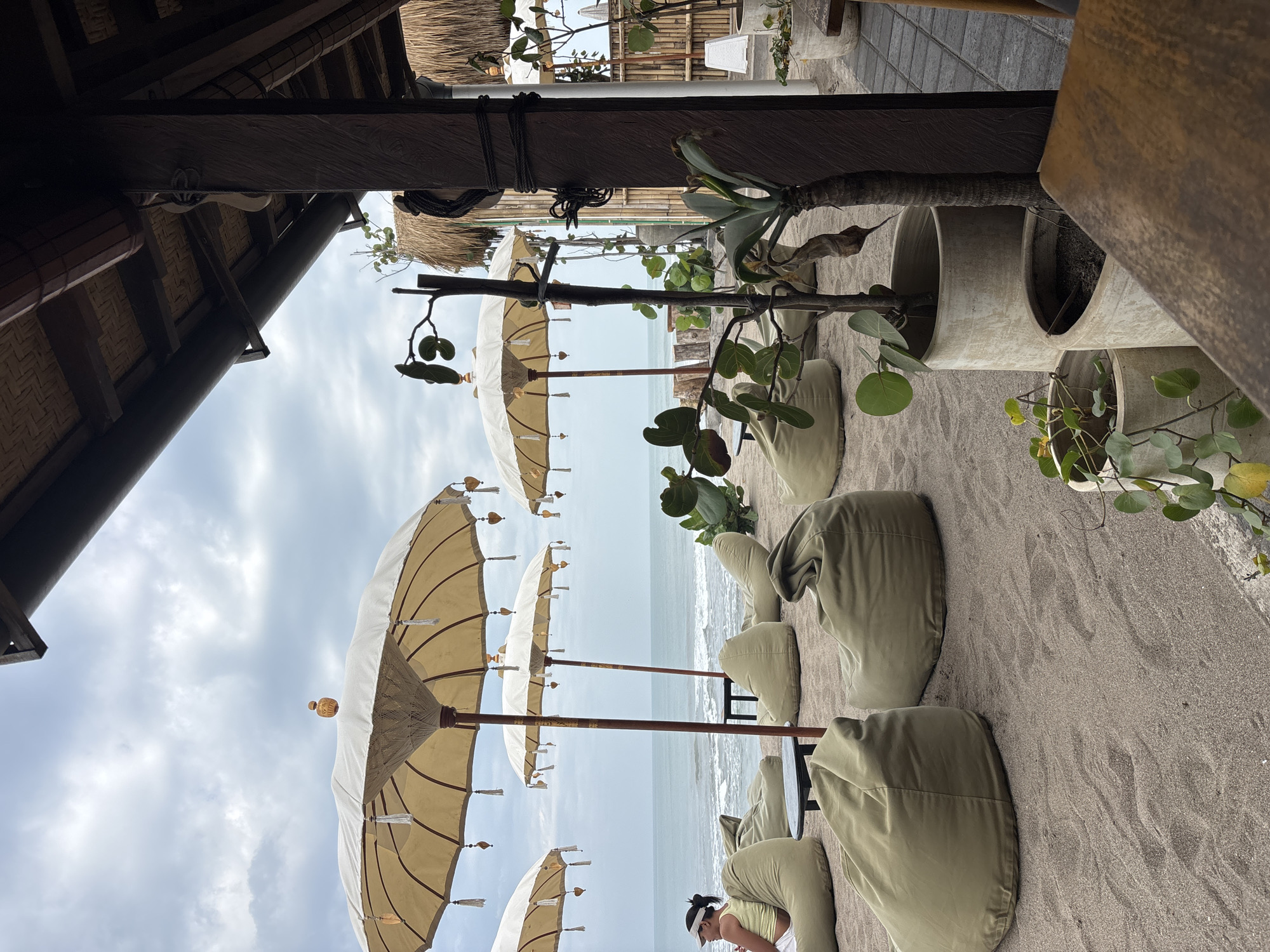 The image depicts a beachside seating area with multiple large, olive-green beanbag chairs arranged on the sand. Several large, light-colored umbrellas with decorative tassels are set up to provide shade. In the background, the ocean is visible with gentle waves under a cloudy sky. A person wearing a yellow top and a white visor is seated on one of the beanbag chairs, facing the ocean. To the right, there is a wooden structure with a thatched roof and bamboo walls, and potted plants with green foliage are placed in front of it. The scene appears to be a relaxed outdoor lounge area at a beach in North Kuta, Indonesia.