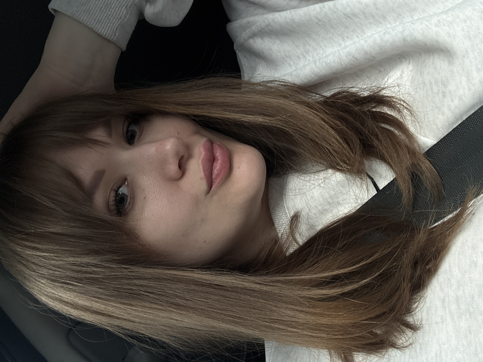 A close-up, rotated selfie shows a woman with shoulder-length brown and blonde highlighted hair. She has fair skin and is wearing a light-colored sweatshirt. A seatbelt strap is visible diagonally across her chest. The location is indeterminable based on the image. No evidence is present to determine if the photo was taken in Chaikovsky, Russia.