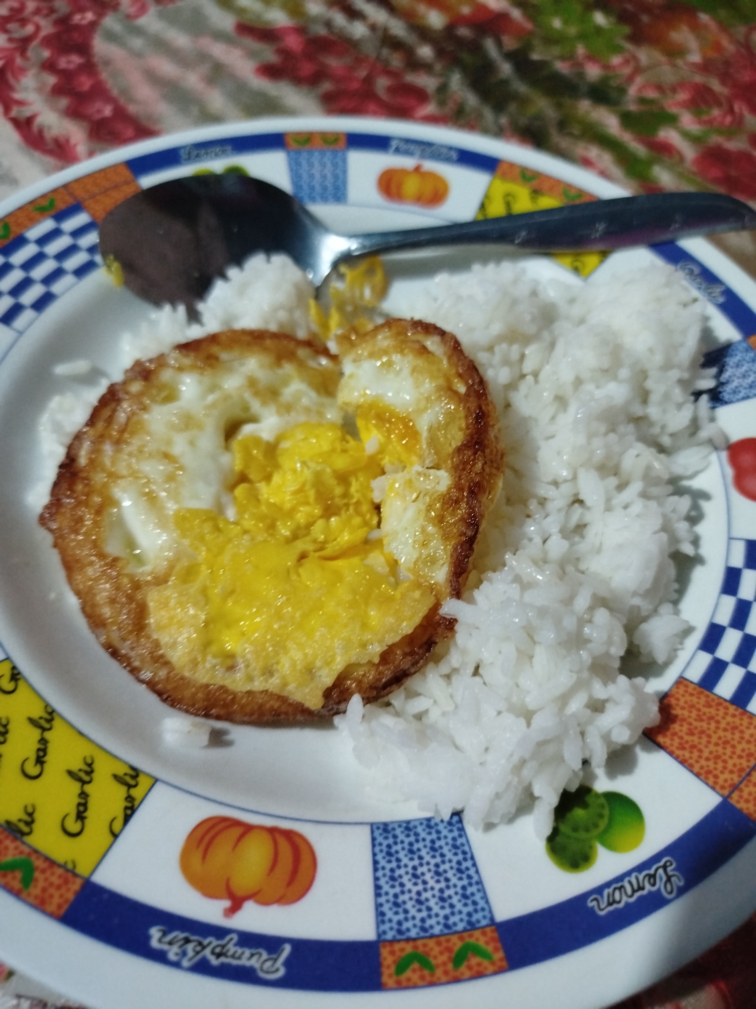 This is a close-up, overhead shot of a white plate with a decorative rim, filled with white rice and a fried egg. The plate's rim features a repeating pattern of colorful squares and circles, with some squares containing images of vegetables and text. One square has a pumpkin icon with the word "Pumpkin" written below it in cursive. Another square has a lime icon with the word "Lemon" written below it in cursive. The food is centered on the plate. A silver spoon rests on the rice, partially covering a portion of the fried egg. The fried egg has a fully cooked white and a runny, bright yellow yolk. The rice appears fluffy and slightly glistening. The background is out of focus, showing a floral-patterned tablecloth in shades of red, pink, and green. This scene is likely from a home kitchen or a local eatery in Balikpapan, Indonesia, given the visual elements and the commonality of such dishes.