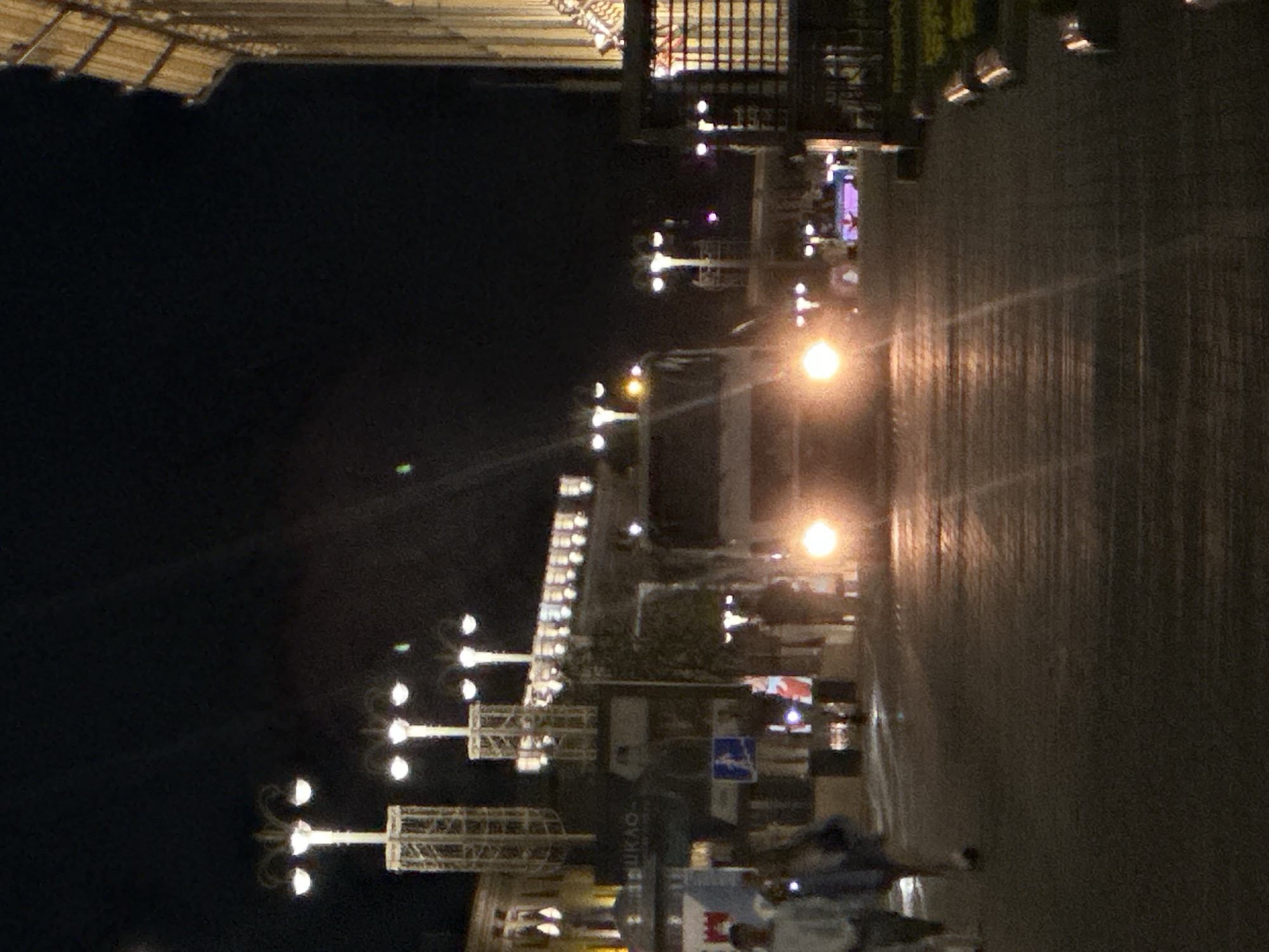 The image shows a street scene at night in an urban environment. A bus with its headlights on is prominent in the center of the frame, driving towards the viewer. Several streetlights with multiple lamps illuminate the scene, casting light on the wet pavement. In the foreground, two people are walking away from the viewer, their figures blurred by the low light conditions. The background features buildings with illuminated windows and decorative structures. A sign with Cyrillic lettering is visible on one of the buildings. The overall atmosphere is that of a busy city street after dark, with artificial lighting creating reflections on the ground.