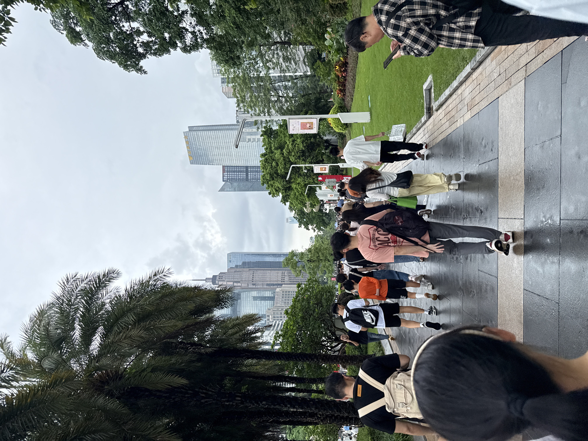 A group of pedestrians walks along a wet, paved pathway in a park-like urban setting. Tall palm trees line the left side of the path, with their fronds reaching towards a cloudy sky. In the background, modern skyscrapers dominate the skyline, indicating a city environment. The people are diverse in age and are dressed in casual clothing, many carrying backpacks or shopping bags. Some individuals are looking at their phones, while others are engaged in conversation as they walk. The pathway itself is made of large, dark stone tiles interspersed with lighter colored ones, showing reflections from the overcast conditions. A manicured green lawn with some colorful foliage runs alongside the right side of the path. The overall scene suggests a public gathering or a leisurely stroll in a metropolitan park. This scene is located in Tianhe District, China.